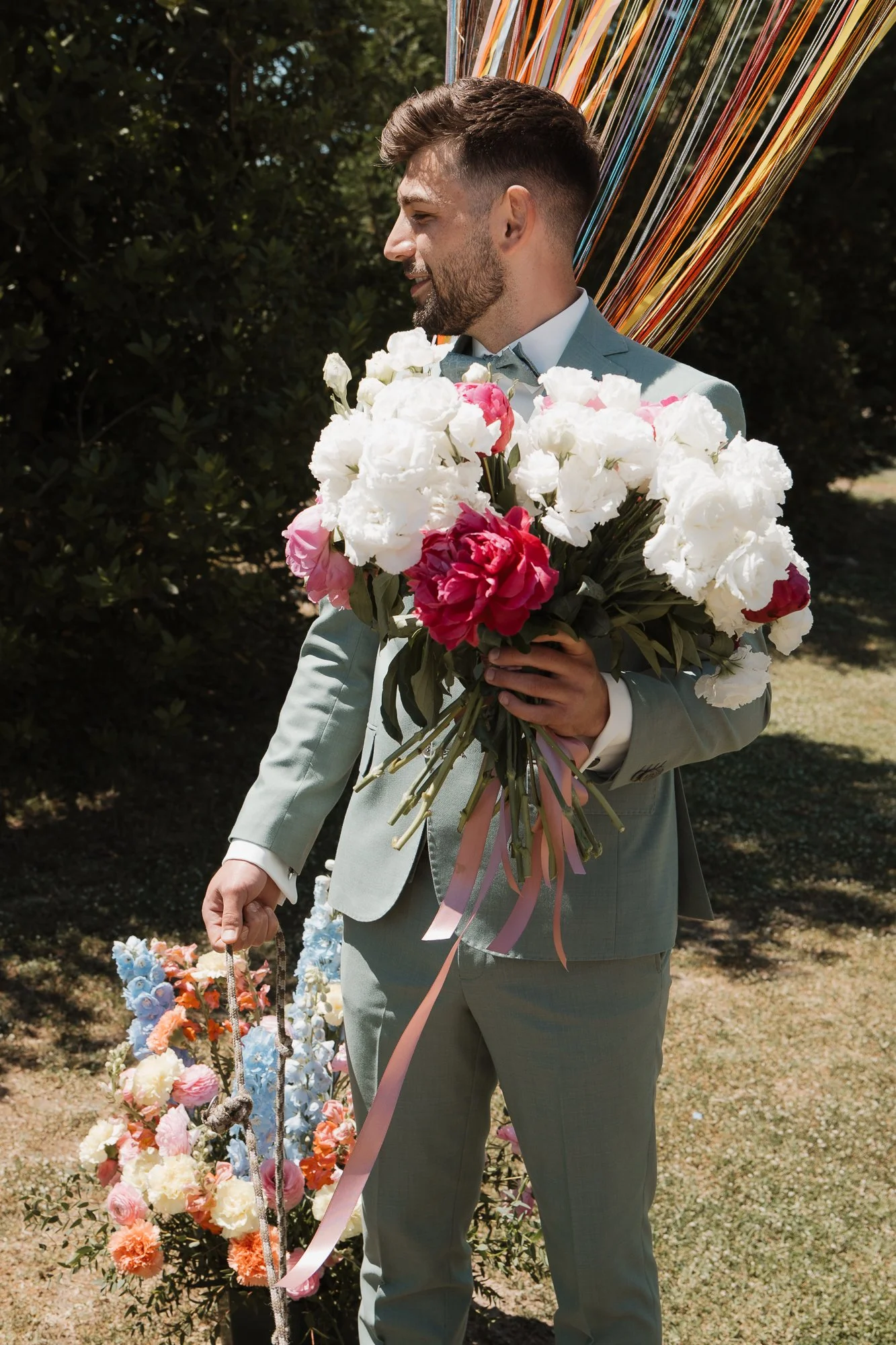 Ein Mann in einem grauen Anzug hält einen großen Blumenstrauß mit weißen und pinkfarbenen Blumen, im Hintergrund sind bunte dekorative Elemente und eine Pflanzenhecke.