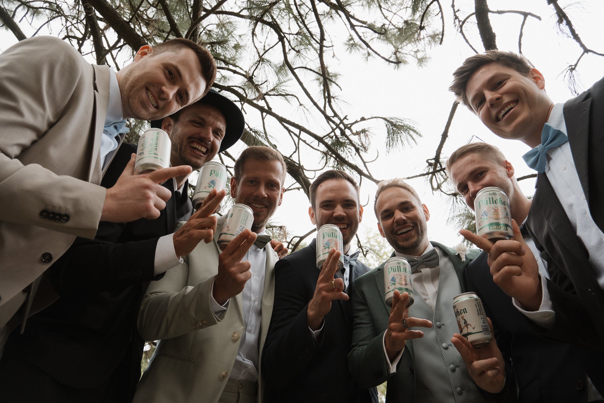 Eine Gruppe von Männern in formeller Kleidung unter einem Baum, die gemeinsam Getränke in Dosen halten und fröhlich in die Kamera lächeln.