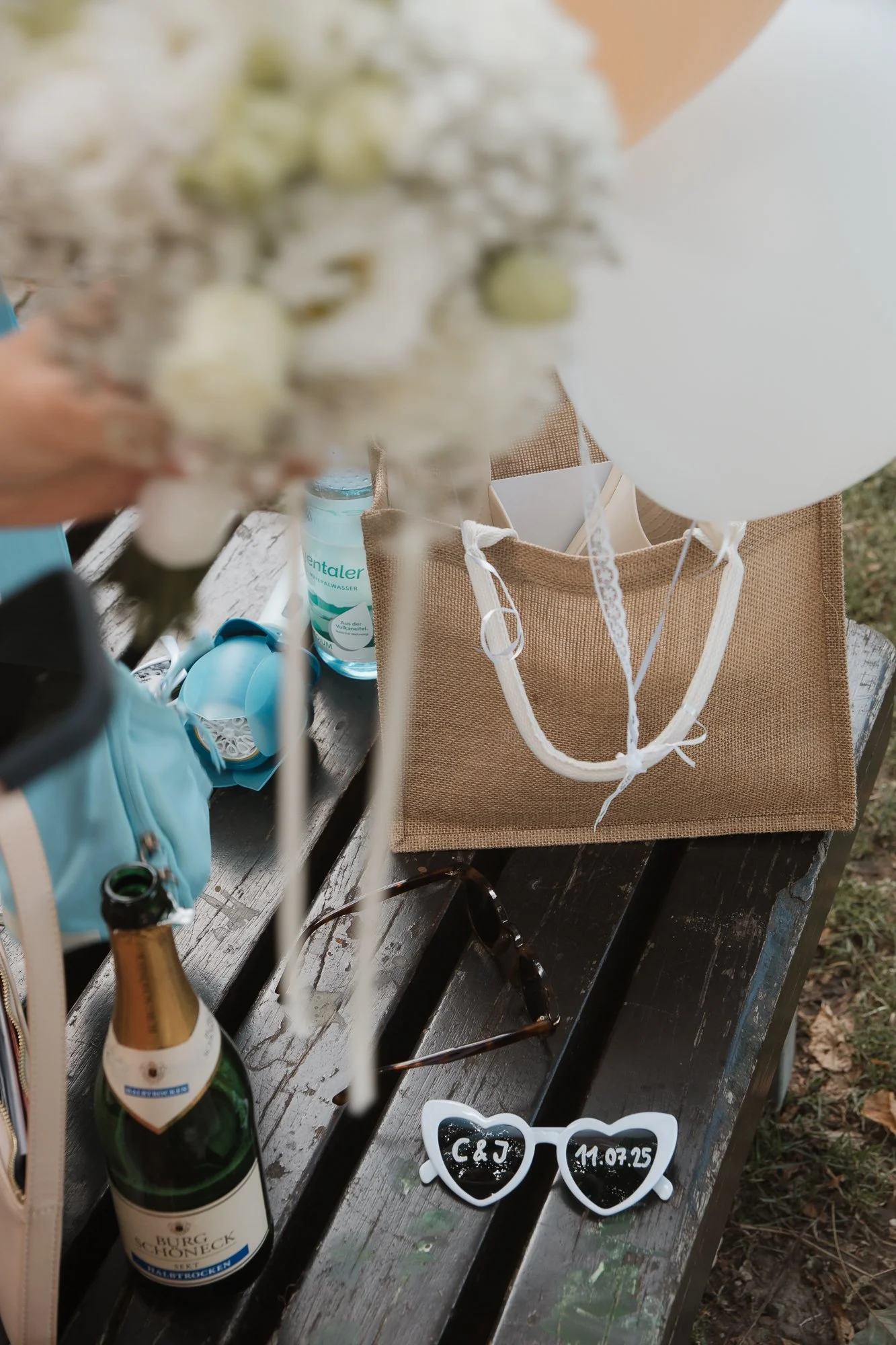 Auf einem dunklen Holzbank liegen Sonnenbrille mit Herzrahmen, eine Flasche Sekt, eine Stofftasche, eine Wasserflasche, eine Brille, eine kleine blaue Tasche, Ballons und eine Flasche Händedesinfektionsmittel. Im Hintergrund sind verschiedene Gegenst