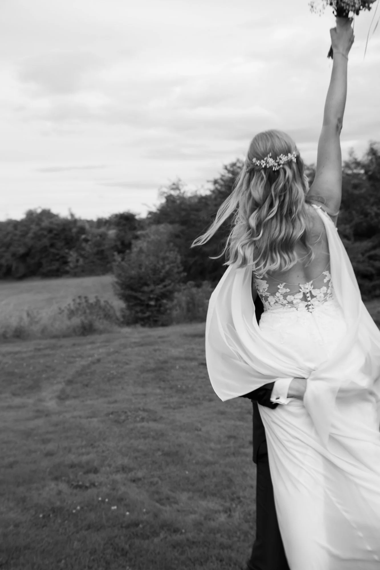 Braut in Hochzeitskleid, die mit ihrem Kleid tanzt, während der Bräutigam sie umarmt, im Freien auf einer Wiese, in Schwarz-Weiß-Fotografie.