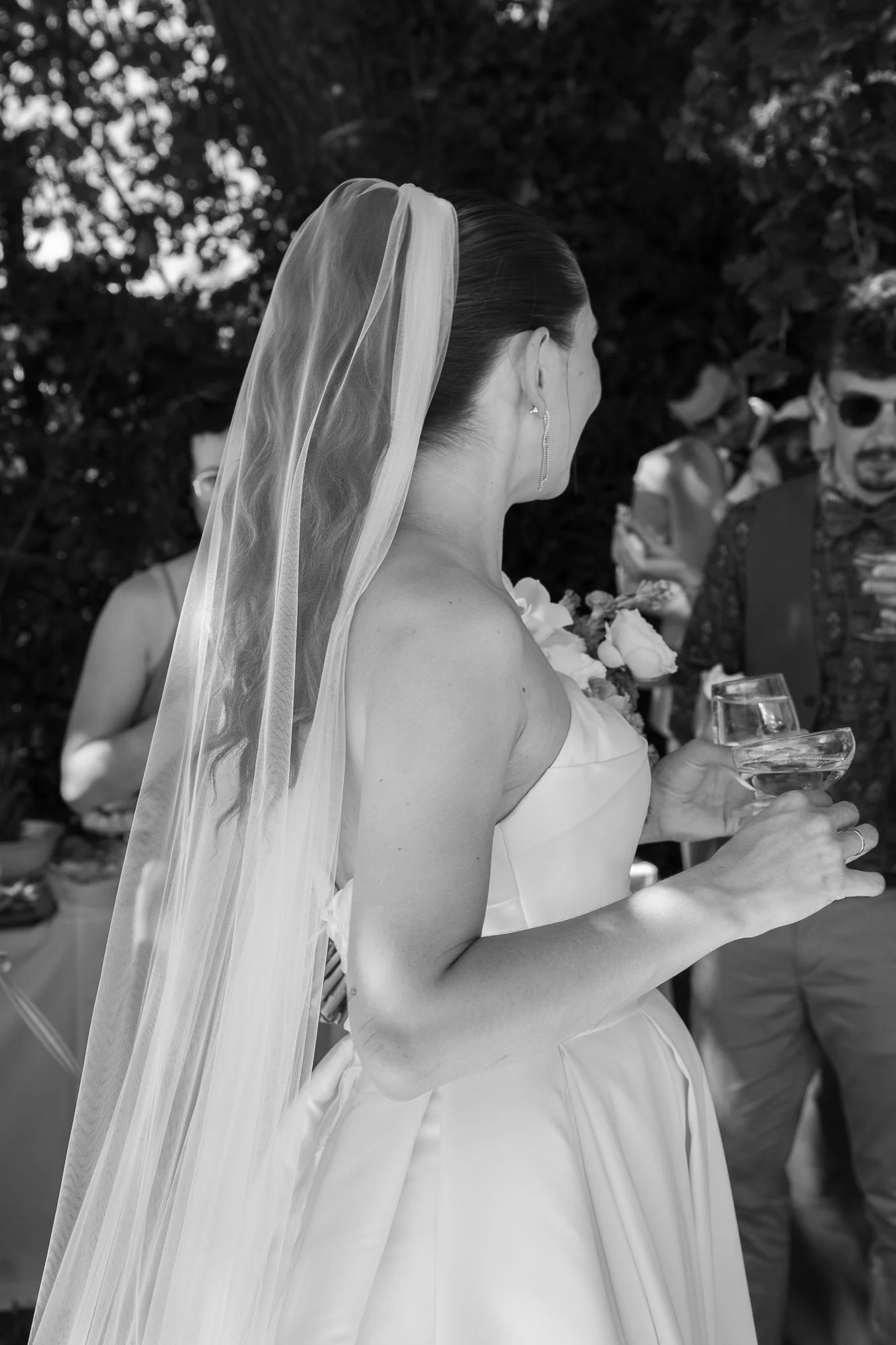 Eine Braut trägt ein Hochzeitskleid und einen Schleier, während sie ein Weinglas hält bei einer Hochzeit im Freien, umgeben von Gästen.