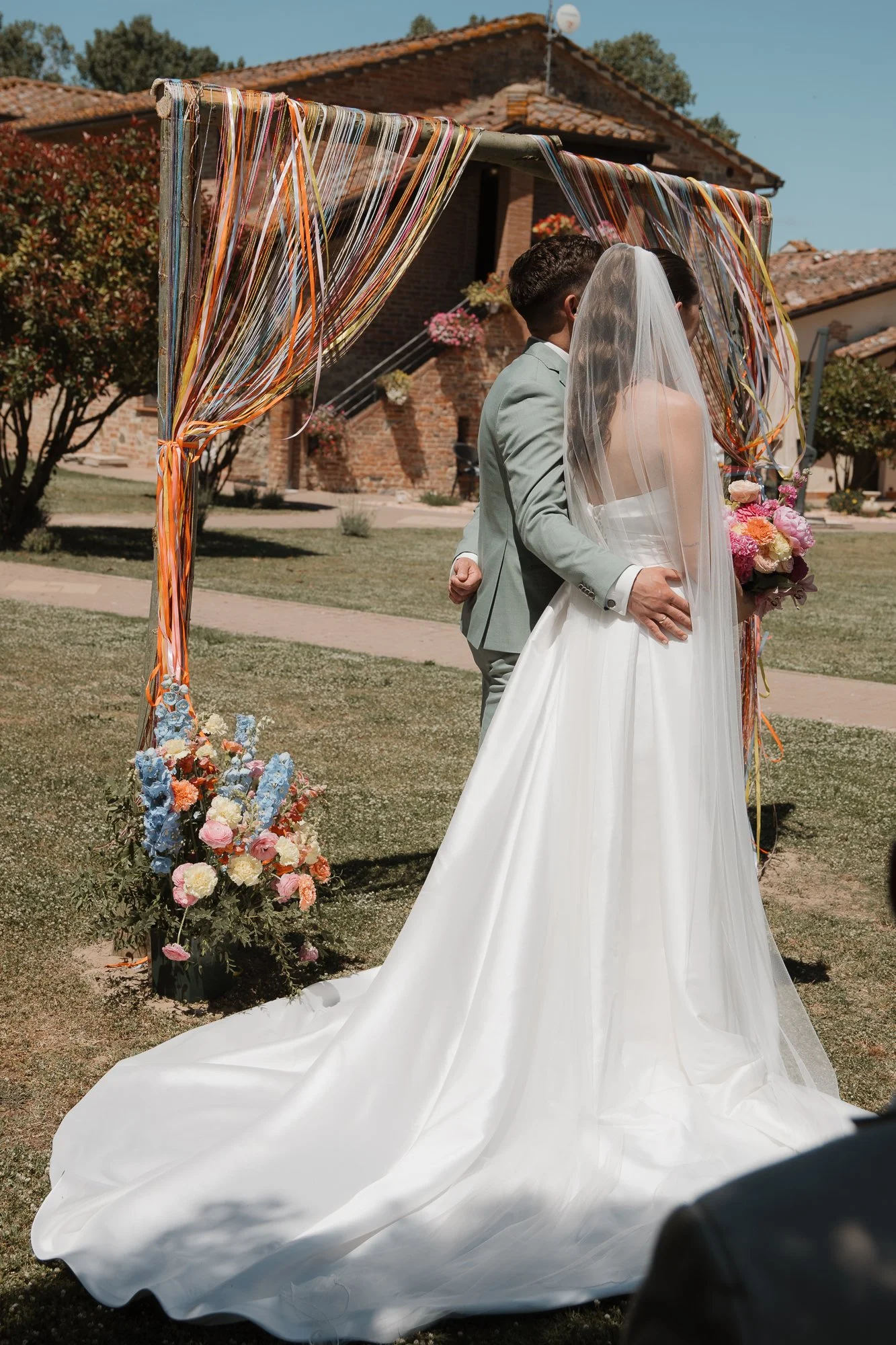 Ein Brautpaar bei einer Trauung im Freien vor einem hölzernen Gewölbe, mit Blumen und bunten Bändern dekoriert.