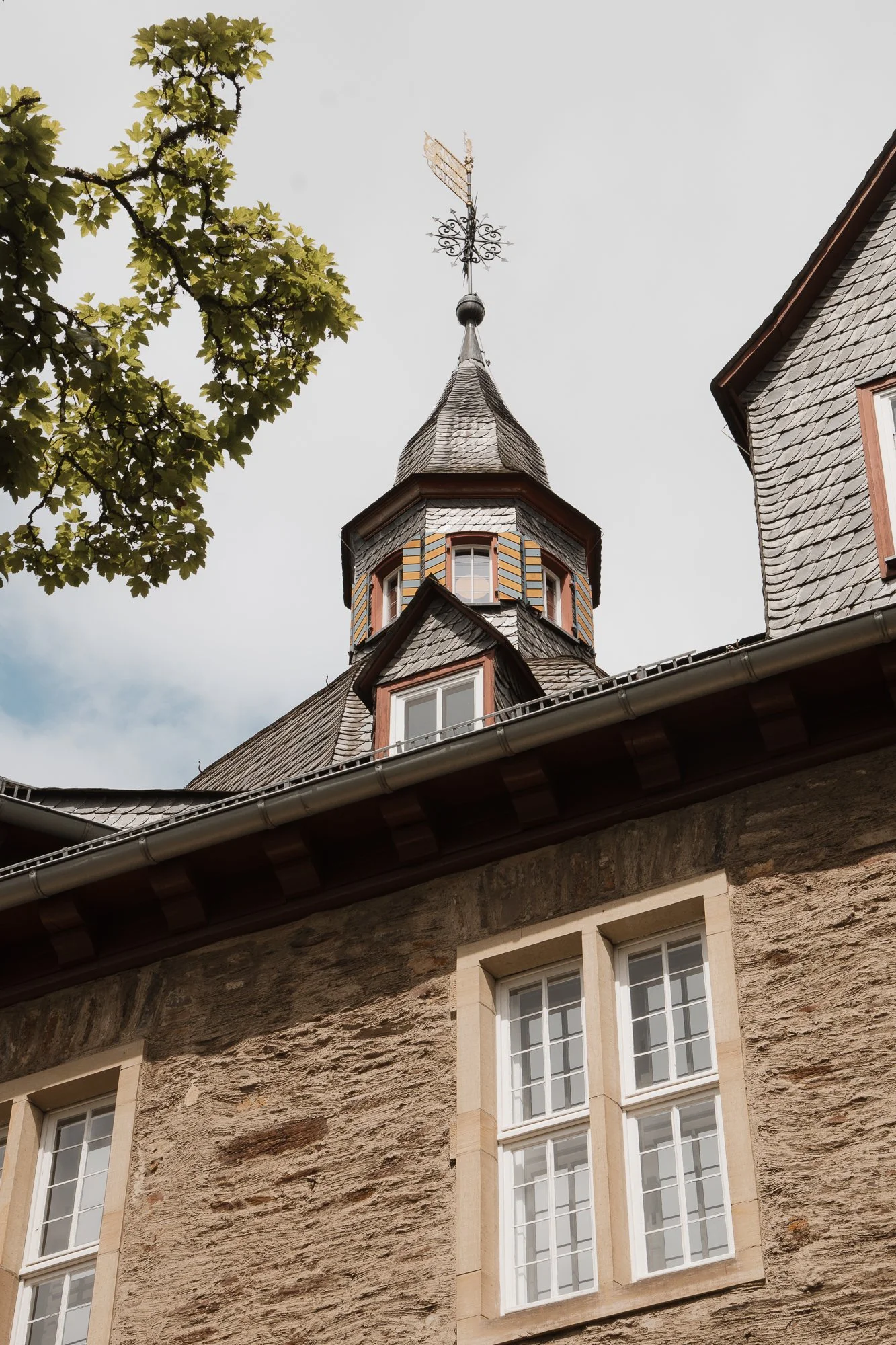 Ein Gebäude mit Steinmauer, weißen Fenstern und einem spitzen Turm mit Schindeln an einem bewölkten Himmel, neben einem Baum.