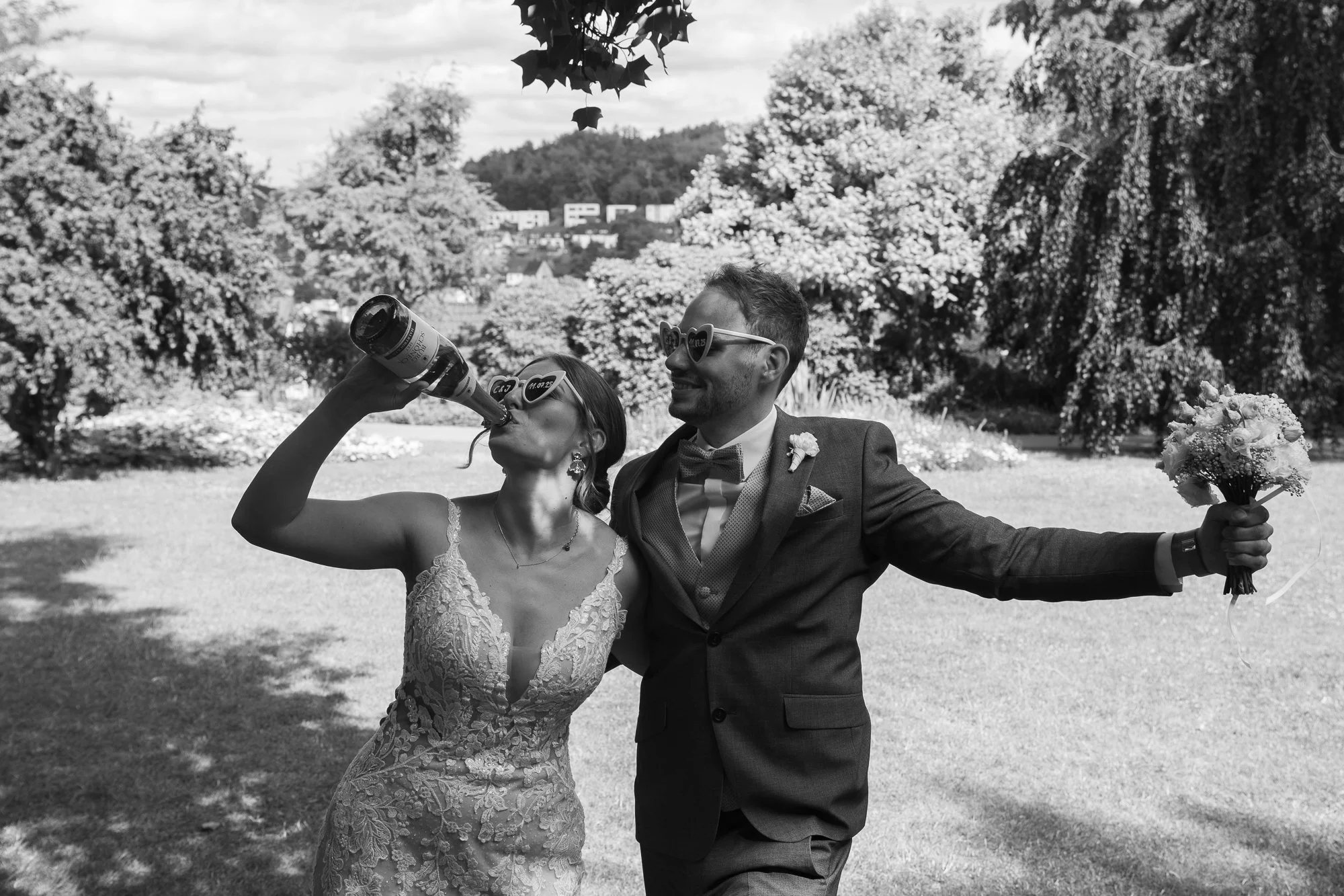 Ein frisch verheiratetes Paar auf einer Wiese, die Braut trinkt aus einer Flasche, während der Bräutigam mit Blumenstrauß in der Hand lächelnd neben ihr steht, umgeben von Bäumen im Freien.