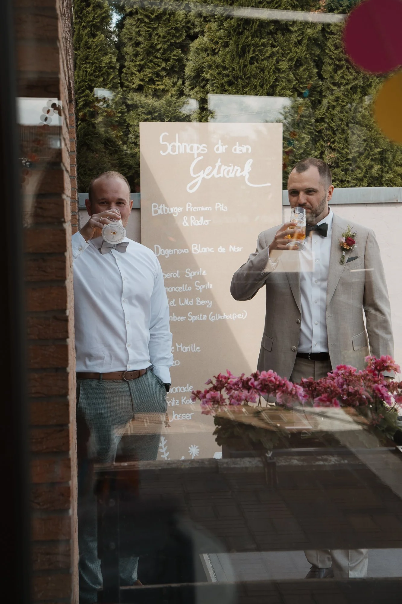 Zwei Männer in formeller Kleidung trinken Bier vor einem Menü-Board mit Getränken, Blick durch eine Glasscheibe