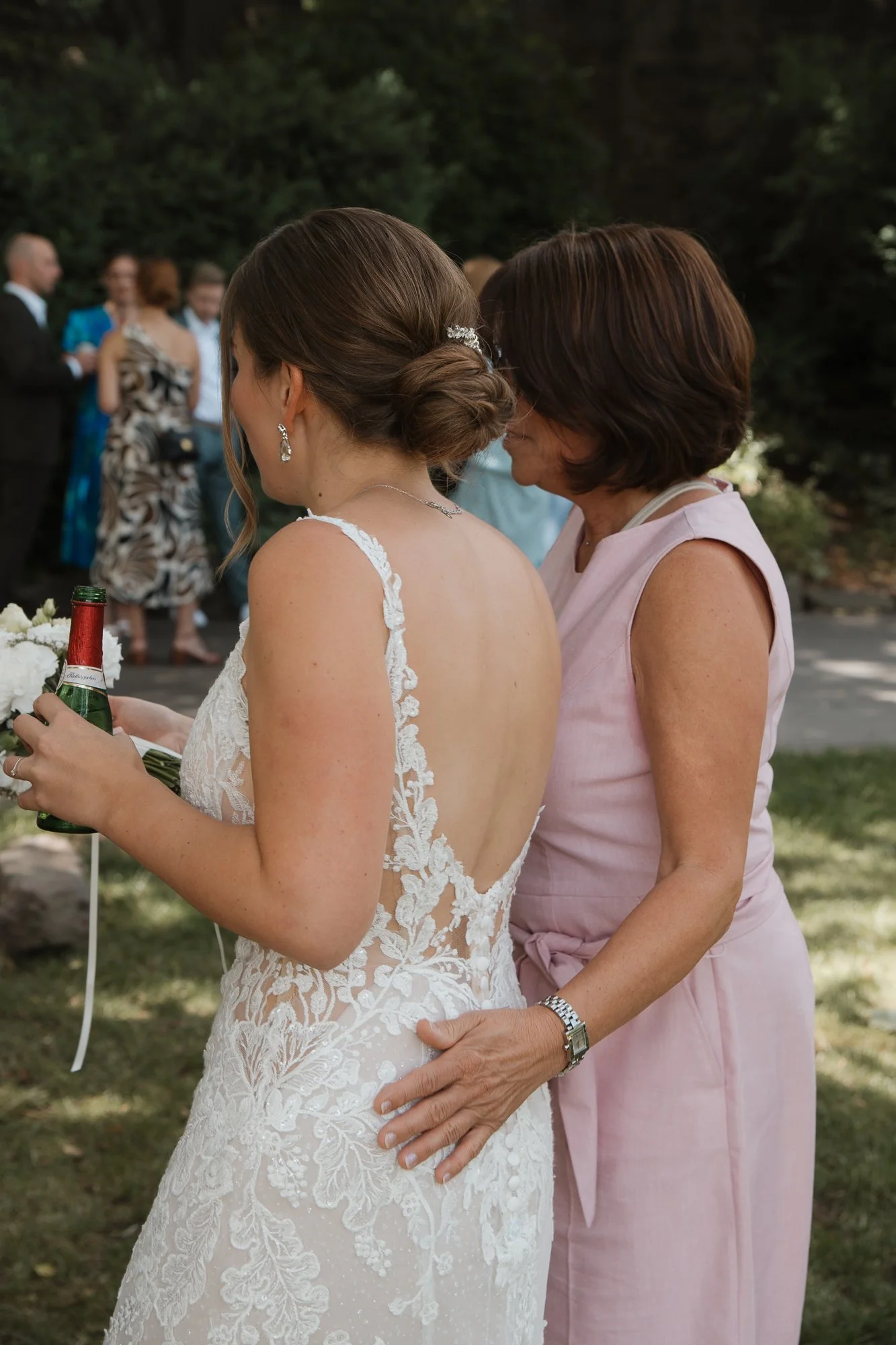 Eine Braut in einem weißen, verzierten Kleid bekommt eine Umarmung von einer älteren Frau bei einer Hochzeit im Freien.