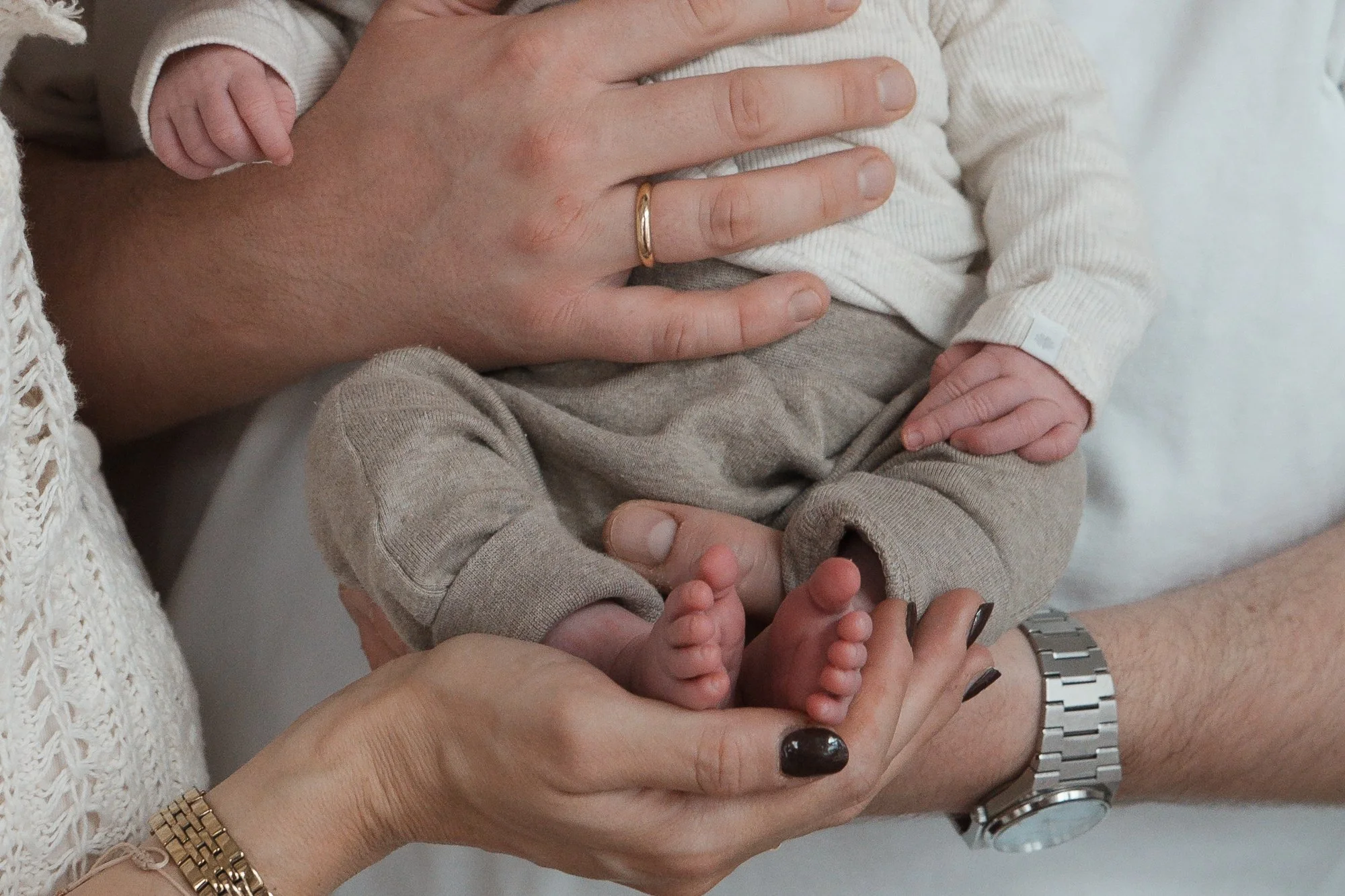 Nahaufnahme von einem Baby, das auf den Händen von zwei Erwachsenen liegt, die es vorsichtig halten. Das Baby trägt einen cremefarbenen Pullover und graue Hosen, die Füße sind sichtbar.