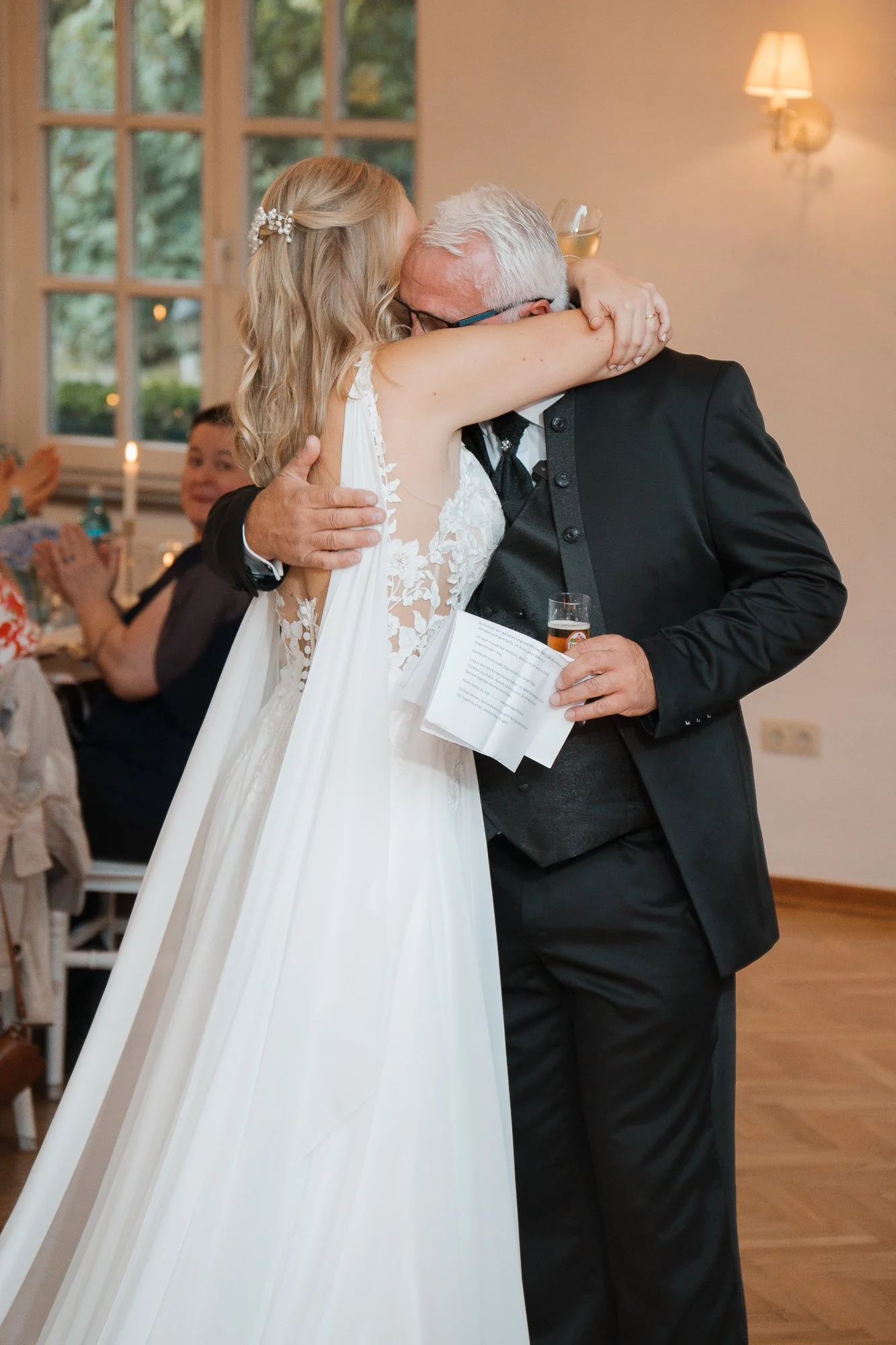 Braut umarmt älteren Mann bei Hochzeit, im Hintergrund feiern Gäste.
