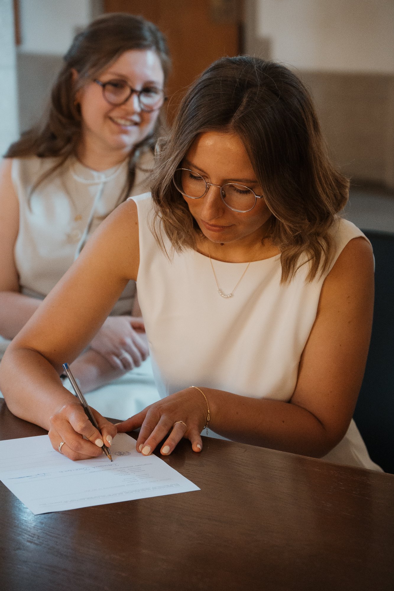 Zwei Frauen beim Unterschreiben eines Dokuments an einem Holztisch, eine sitzt und schreibt, die andere steht im Hintergrund, lächelnd.