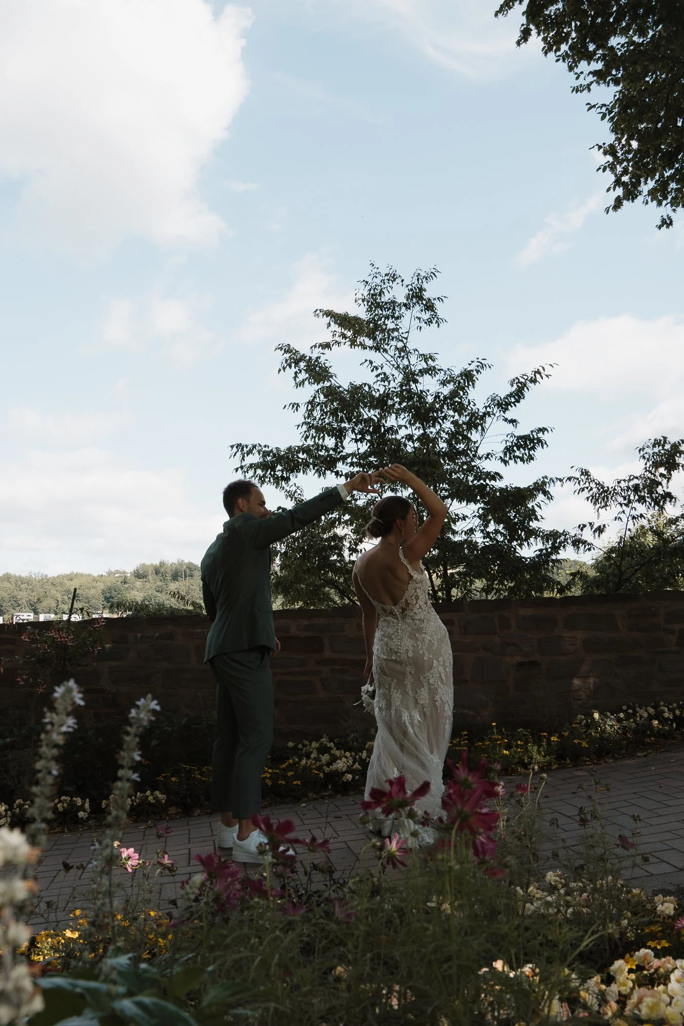 Ein Paar tanzt im Freien, die Frau trägt ein weißes Hochzeitskleid und der Mann einen Anzug, vor einer Mauer mit Bäumen im Hintergrund.