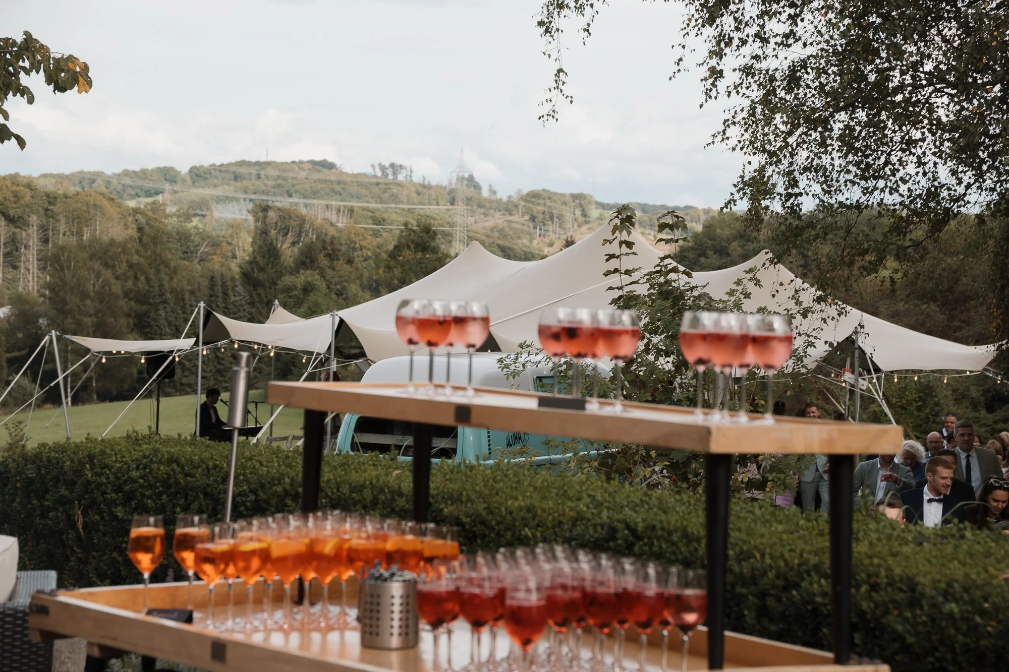 Selbstbedienung mit rosafarbenen Getränken in Weingläsern bei einer Outdoor-Party, im Hintergrund große weiße Zelte und Menschen in formeller Kleidung