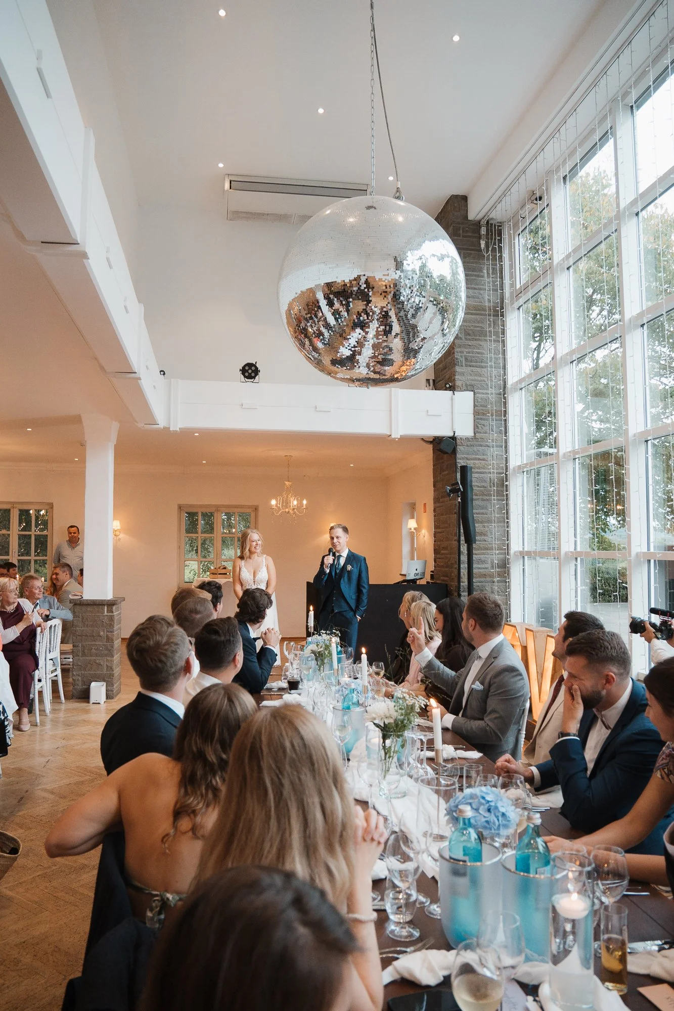 Hochzeitsfeier in einem festlich dekorierten Raum, mit Braut und Bräutigam beim Reden, umgeben von Gästen, großer Decken-Spiegel und große Fenster