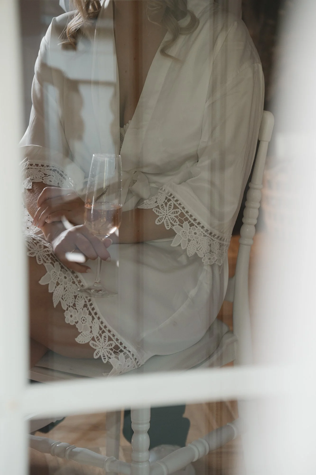 Frau in einem weißen Kleid mit Spitze an den Ärmeln, sitzt auf einem Stuhl, hält ein Weinglas mit Roséwein, gesehen durch eine Fensterscheibe.
