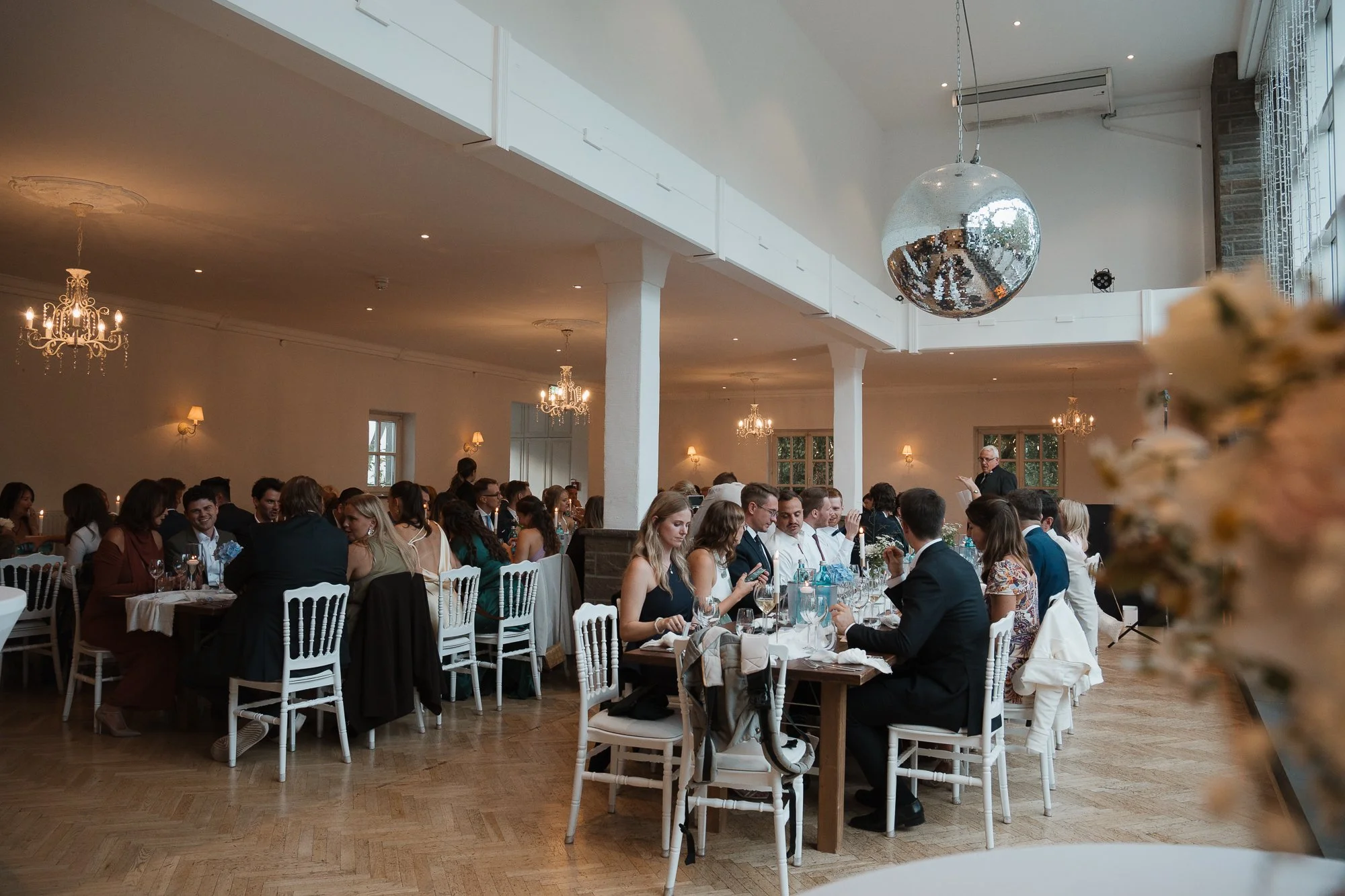Menschen sitzen an mehreren Tischen in einem festlich geschmückten Saal bei einem Event oder einer Feier, während im Hintergrund eine Person spricht.