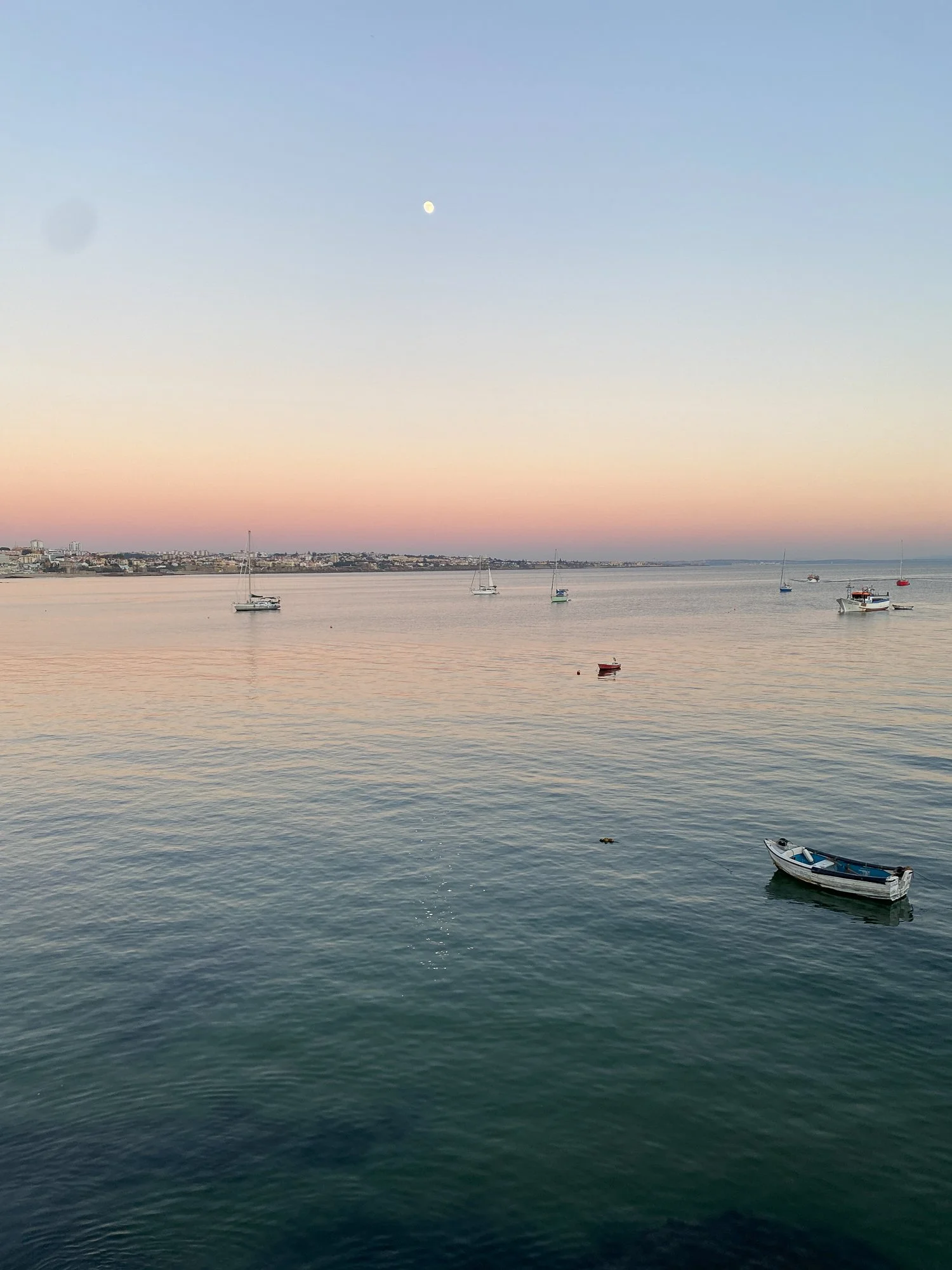 Blick auf einen ruhigen Hafen bei Sonnenuntergang mit mehreren Segelbooten und einem kleinen Motorboot im Wasser, darunter der Mond am Himmel.
