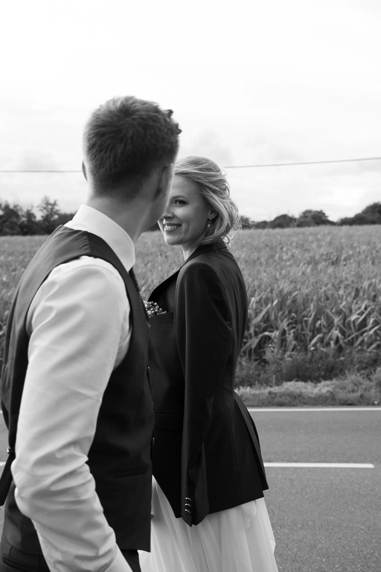 Zwei Menschen, ein Mann und eine Frau, stehen auf einer Straße neben einem Feld. Die Frau lächelt in die Kamera, der Mann steht mit dem Rücken zur Kamera.