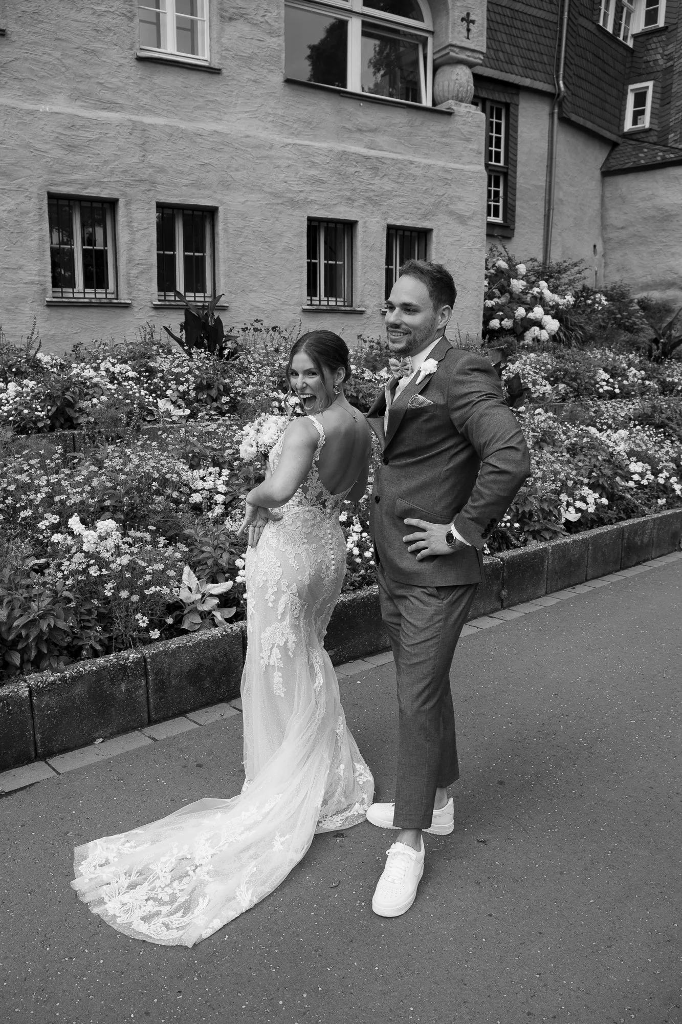 Ein Hochzeitspaar, der Braut in einem eleganten, Lace-Kleid, der Bräutigam in einem Anzug mit Turnschuhen, posieren lachend vor einer Gebäudefassade mit Blumenbeeten.