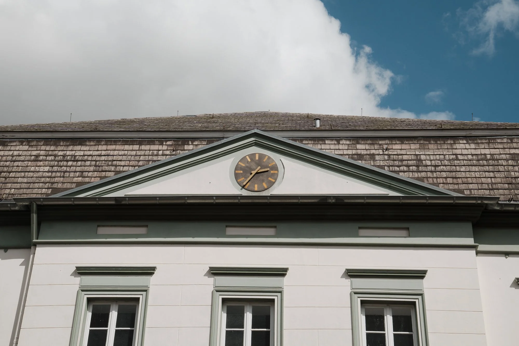 Schulgebäude mit einer Uhr auf dem Dach, weißen Wänden und drei Fenster darunter, Himmel mit Wolken im Hintergrund.