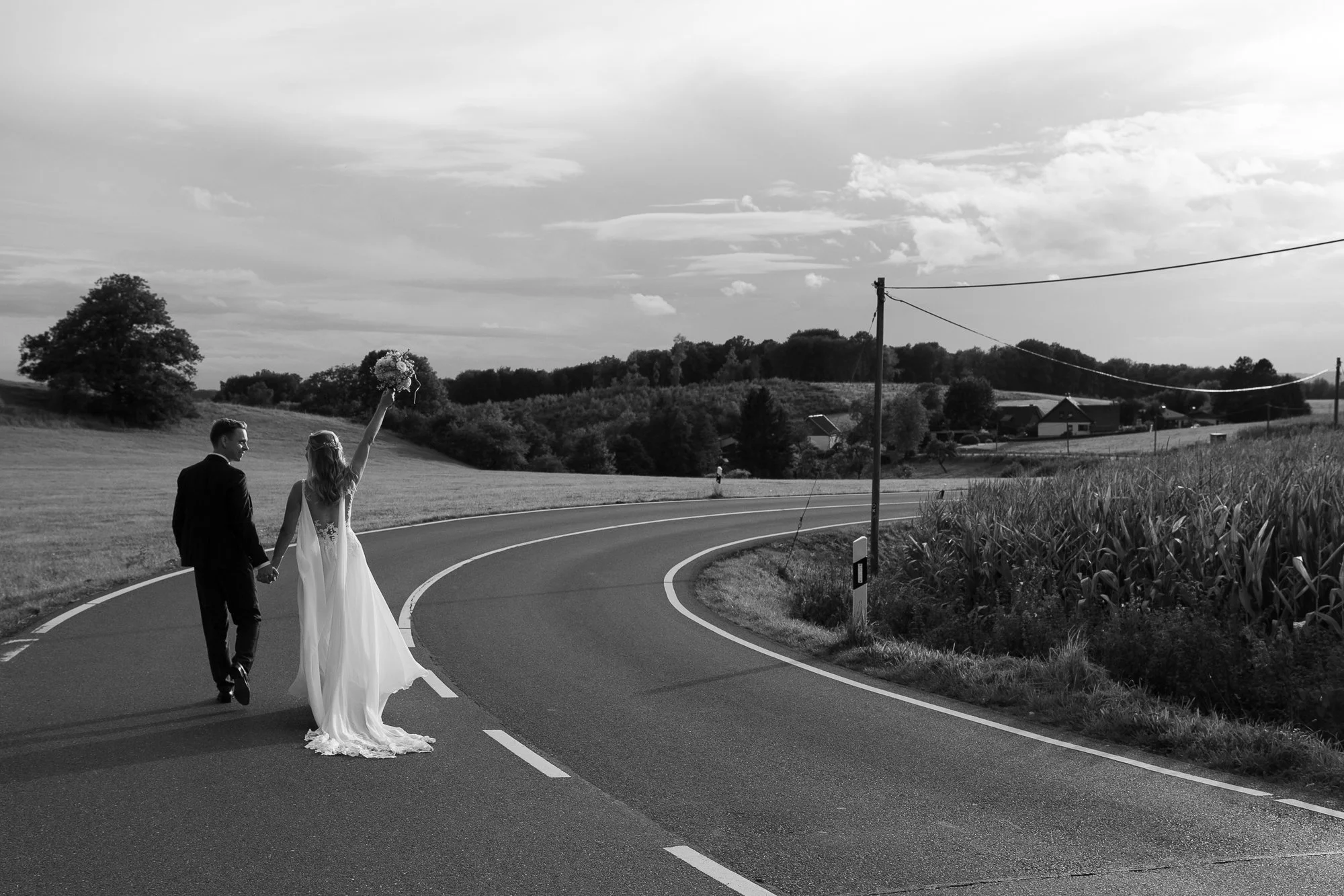 Ein Brautpaar läuft Hand in Hand auf einer kurvigen Landstraße, die Braut hält den Brautstrauß hoch, während sie zum Sonnenuntergang spazieren. Es ist schwarz-weiß.