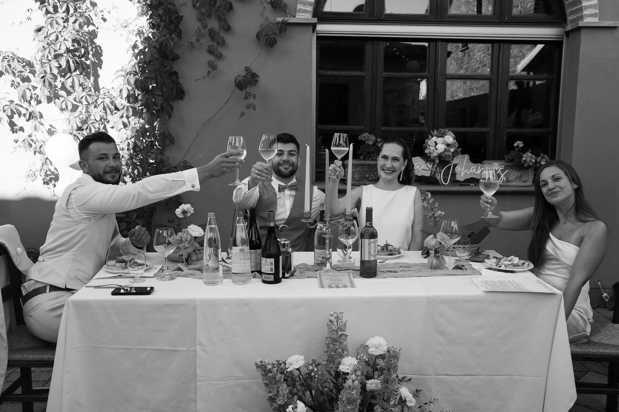 Fünf Menschen sitzen an einem festlich gedeckten Tisch, trinken Wein und lächeln. Der Tisch ist mit Weinflaschen, Gläsern, Blumen und Kerzen geschmückt. Es scheint ein festlicher Anlass wie eine Feier zu sein, wahrscheinlich eine Hochzeit.