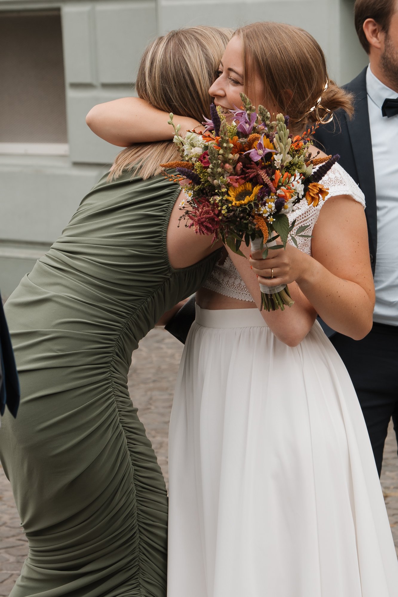 Eine Frau in weißem Brautkleid erhält einen herzlichen Kuss und Umarmung von einer anderen Frau, die ein grünes Kleid trägt, während sie ein Blumenbouquet hält.
