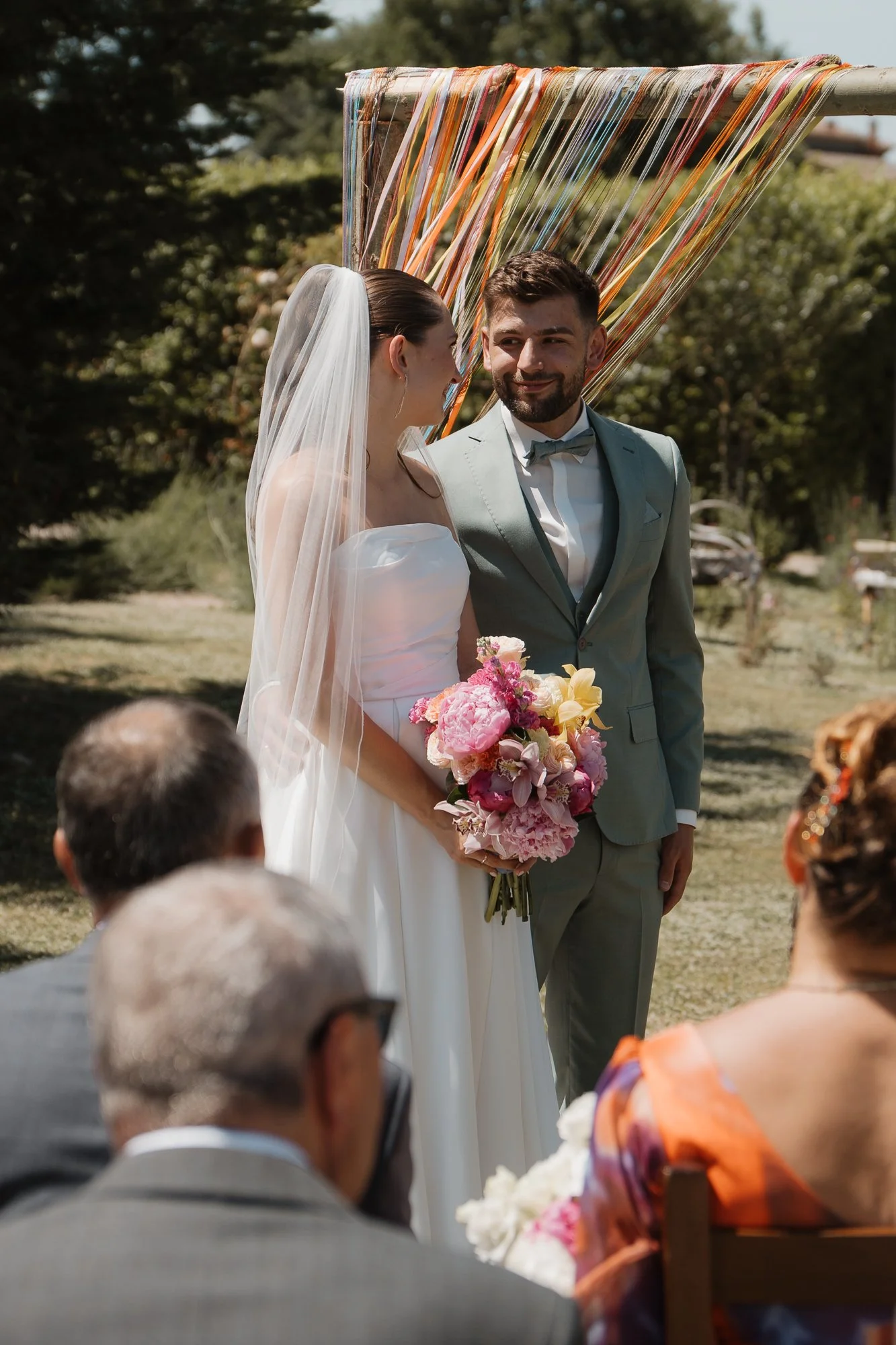 Ein Brautpaar bei einer Trauung im Freien, die Braut trägt ein weißes Kleid und einen Schleier, während der Bräutigam einen hellgrünen Anzug mit Fliege trägt. Die Braut hält einen bunten Blumenstrauß. Es sind Gäste im Vordergrund zu sehen, die der Ze