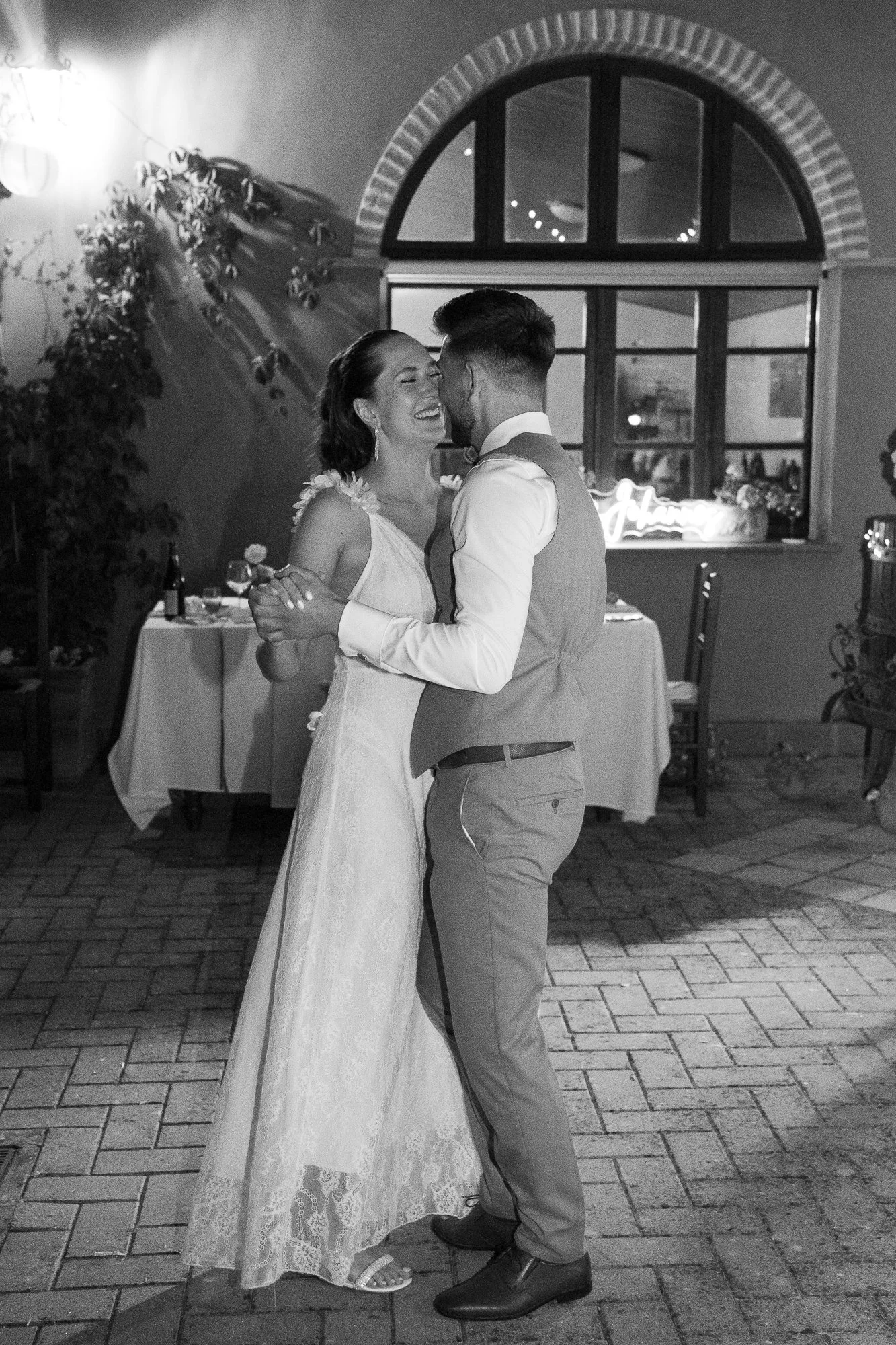 Ein Paar tanzt auf einer Hochzeit, lächelnd und in Umarmung, in einem festlich dekorierten Raum bei Nacht.