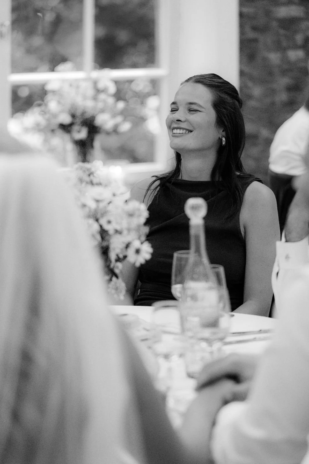Eine lachende Frau mit dunklem Haar bei einer Veranstaltung, im Hintergrund ein Fenster und Blumen. Black-and-white-Foto.
