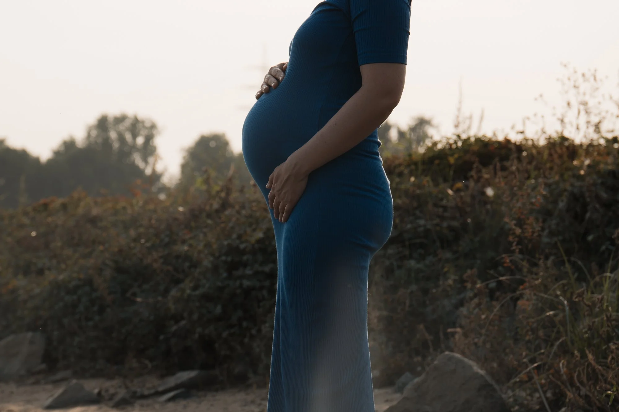 Eine schwangere Frau in einem blauen Kleid steht im Freien, gehaltene Hand auf ihrem Bauch, im Hintergrund Bäume und ein Himmel bei Sonnenuntergang.