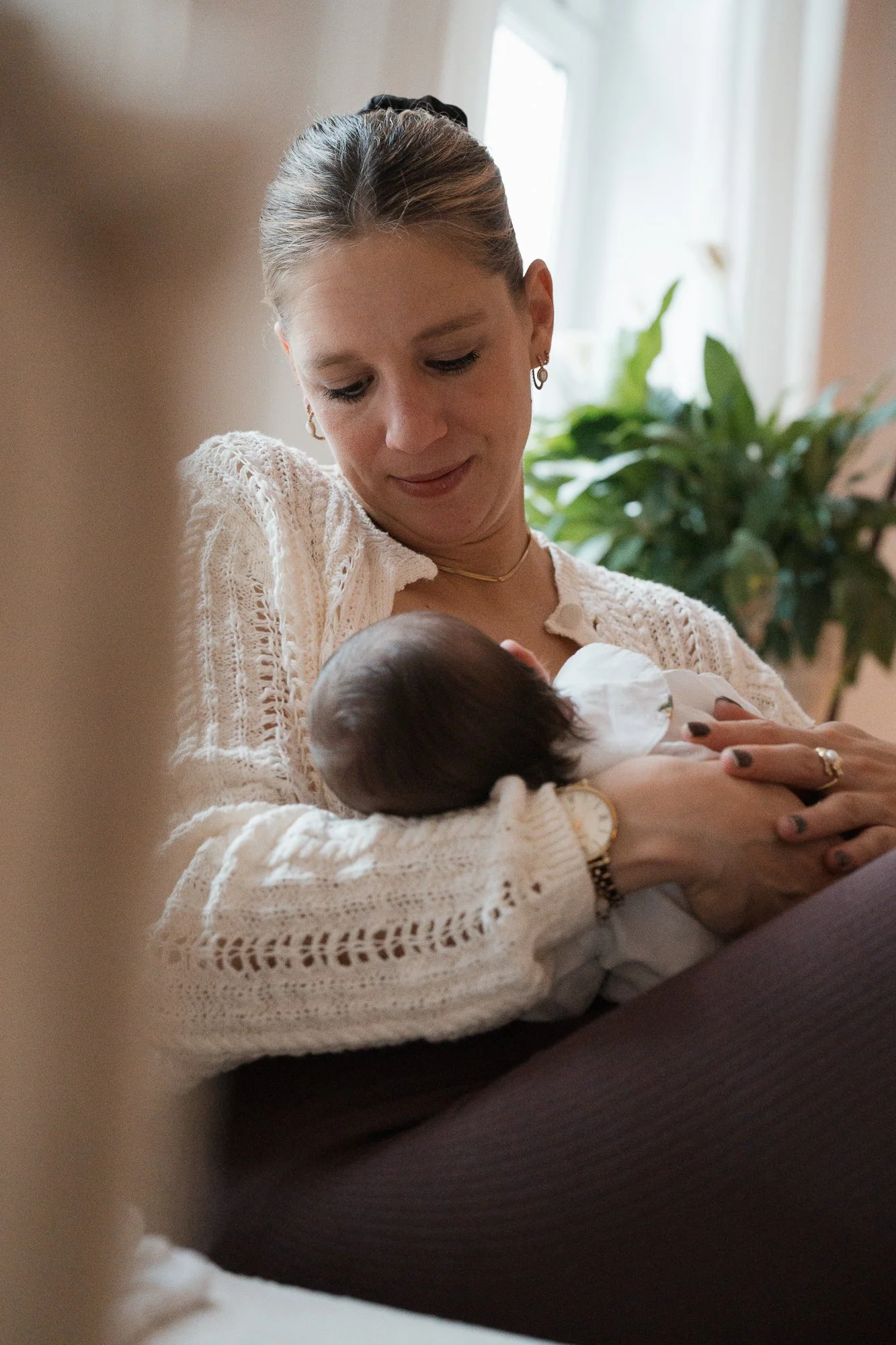 Eine Frau hält ein neugeborenes Baby im Arm und blickt liebevoll auf das Kind. Im Hintergrund sind Pflanzen und ein Fenster sichtbar.