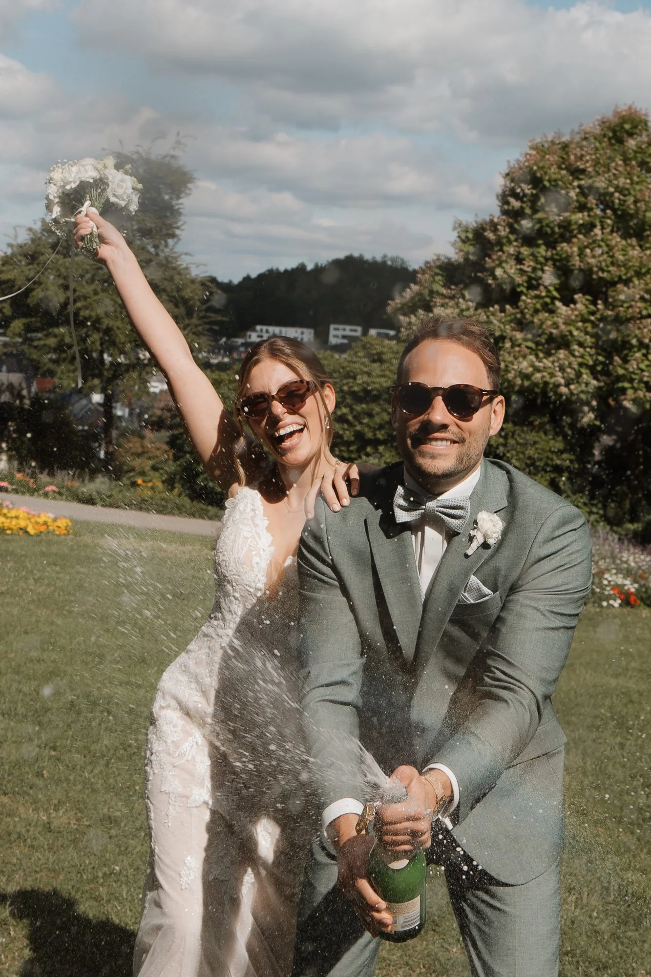Ein Brautpaar feiert draußen, die Frau sprüht mit einem Sektflasche Wasser, beide tragen Sonnenbrillen, im Hintergrund Bäume und ein bewölkter Himmel.