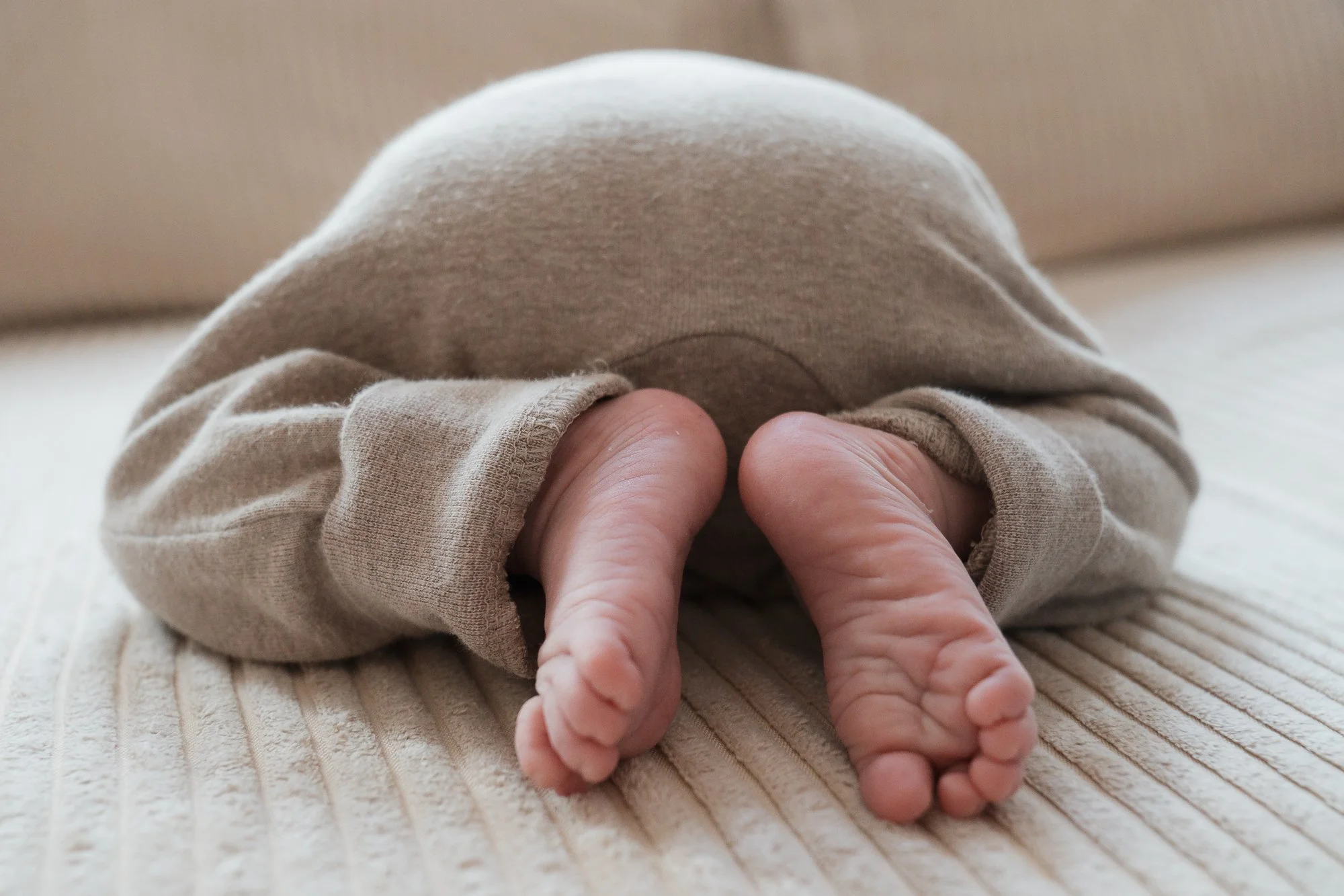 Nahaufnahme von Babyfüßen, die aus einem beige Sweatshirt herausschauen, auf einem gerippten Kissen.