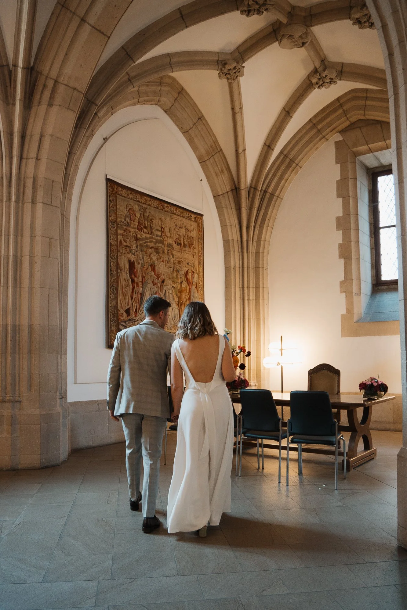 Ein Brautpaar in einem historischen Kirchenraum, das die Hand hält, mit einem Wandteppich und einem Fenster im Hintergrund.