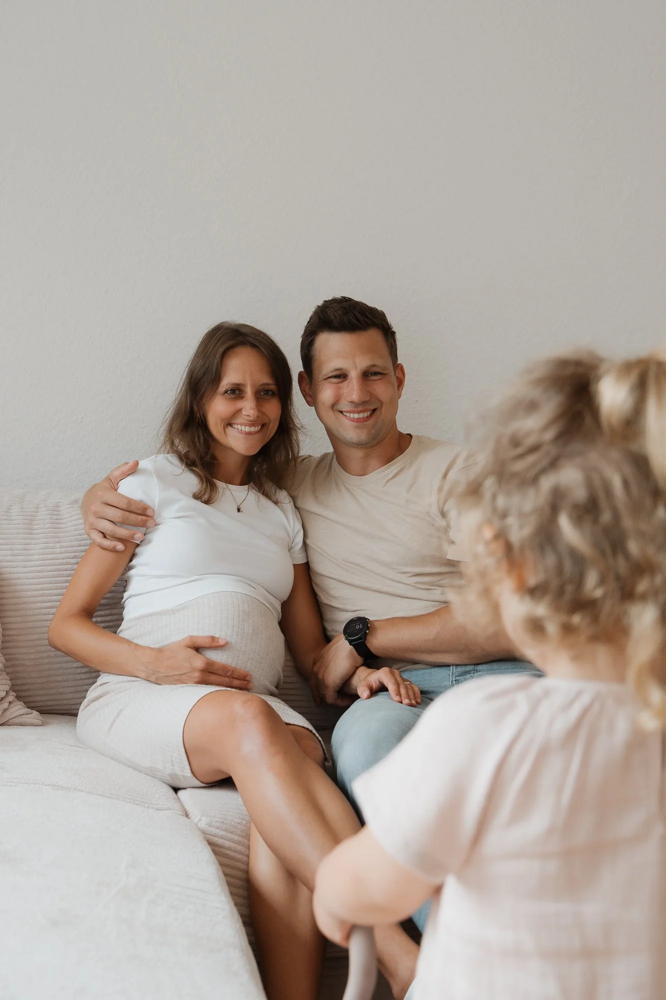Ein lächelndes Paar sitzt auf einem Sofa, die schwangere Frau umarmt den Mann, sie sprechen mit einem Kind mit blonden Locken, das vor ihnen sitzt.