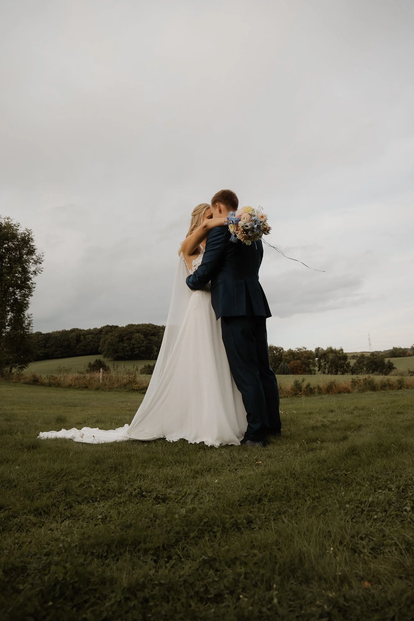 Ein Brautpaar umarmt sich im Freien auf einer grünen Wiese, der Himmel ist bewölkt, die Braut trägt ein weißes Kleid und der Bräutigam einen dunklen Anzug, die Braut hält einen Blumenstrauß.