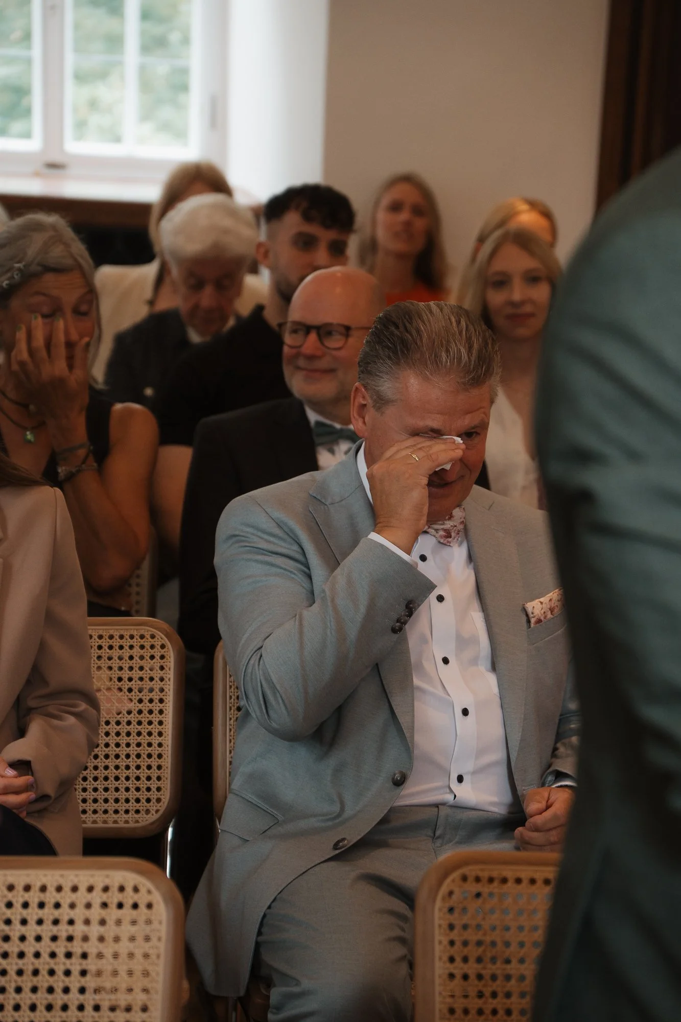 Mehrere Menschen sitzen in einem Raum, die meisten sind lächelnd oder emotional berührt, während eine Person in einem grauen Anzug offensichtlich tränt oder versucht zu schlucken.