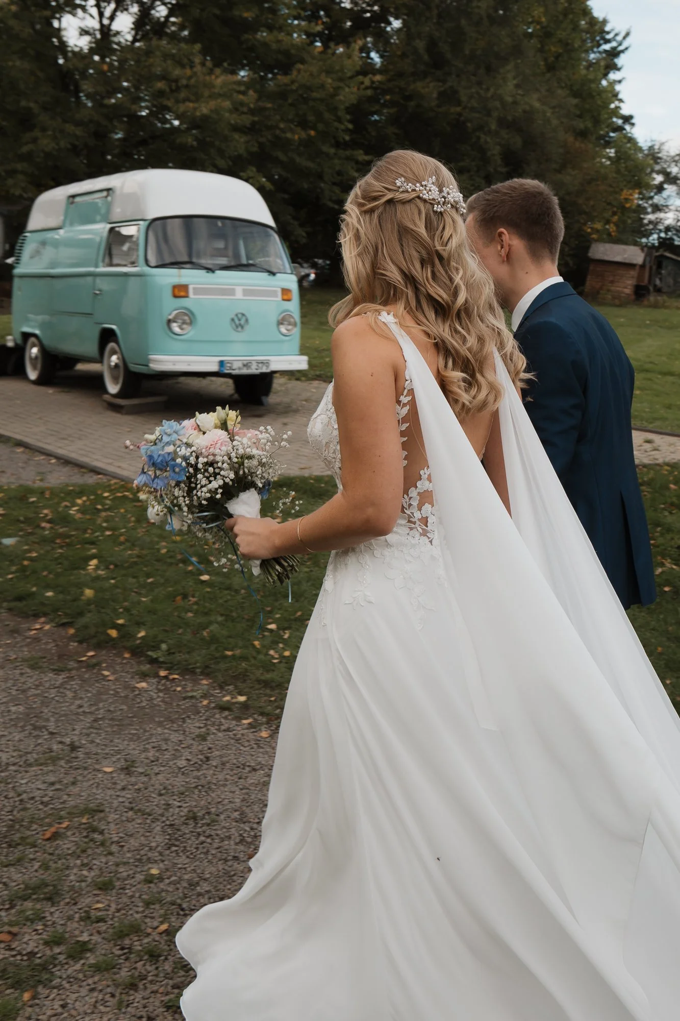 Ein Brautpaar, die Braut hält einen Blumenstrauß, bei sonnigem Wetter, vor einem blauen VW-Bus, im Freien.