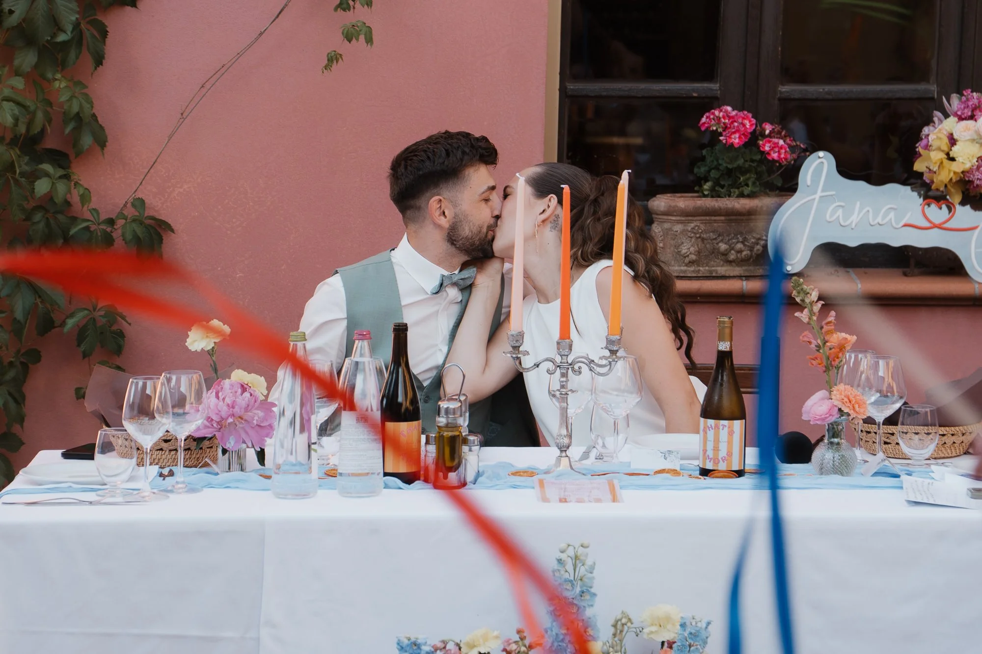 Ein junges Paar küsst sich an einem festlich gedeckten Tisch während einer Hochzeit, mit Blumen und Kerzen als Dekoration.