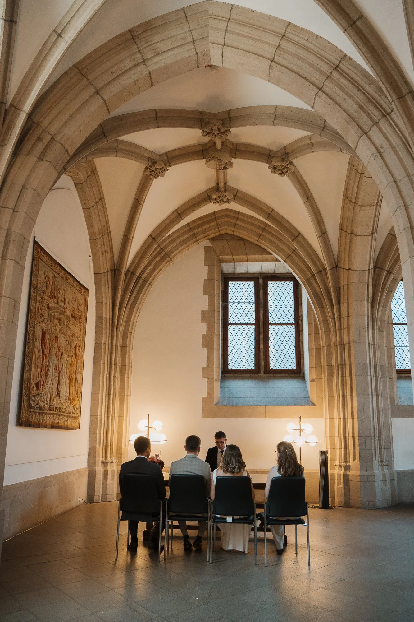 Eine Hochzeitszeremonie in einem gotischen Raum mit Bogenfenstern, bei der fünf Personen, darunter Braut und Bräutigam, an einem Tisch sitzen.