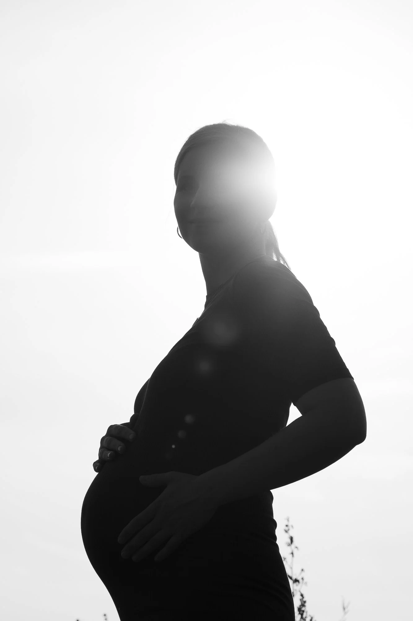 Schwarz-weiß-Foto einer schwangeren Frau im Profil, die vor hellen Himmel steht, mit der Hand auf dem Bauch und leichter Locke in den Haaren.