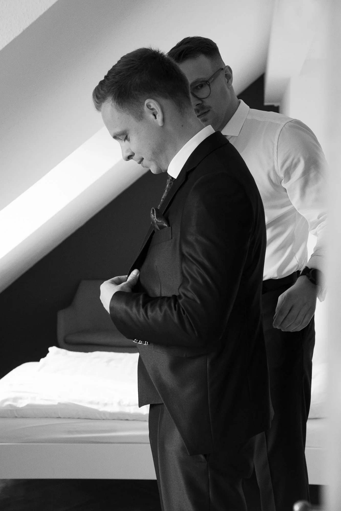 Zwei Männer in formeller Kleidung, einer in einem schwarzen Anzug und der andere in einem weißen Hemd, stehen im Raum.