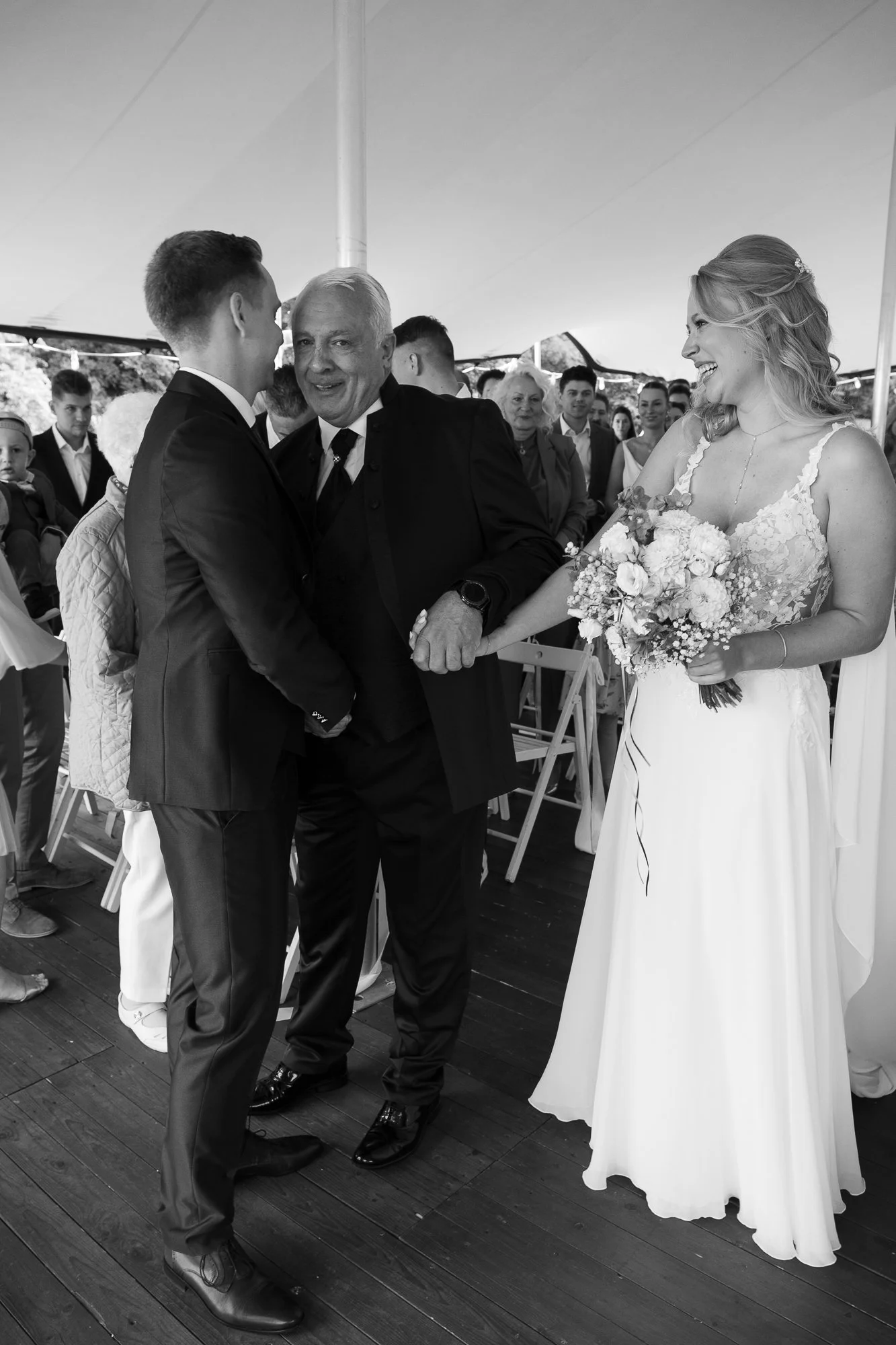 Ein Hochzeitsfoto mit einer lachenden Braut, einem Bräutigam und einem älteren Mann, die sich die Hand geben, unter einem Zelt während einer Hochzeitsfeier im Freien.