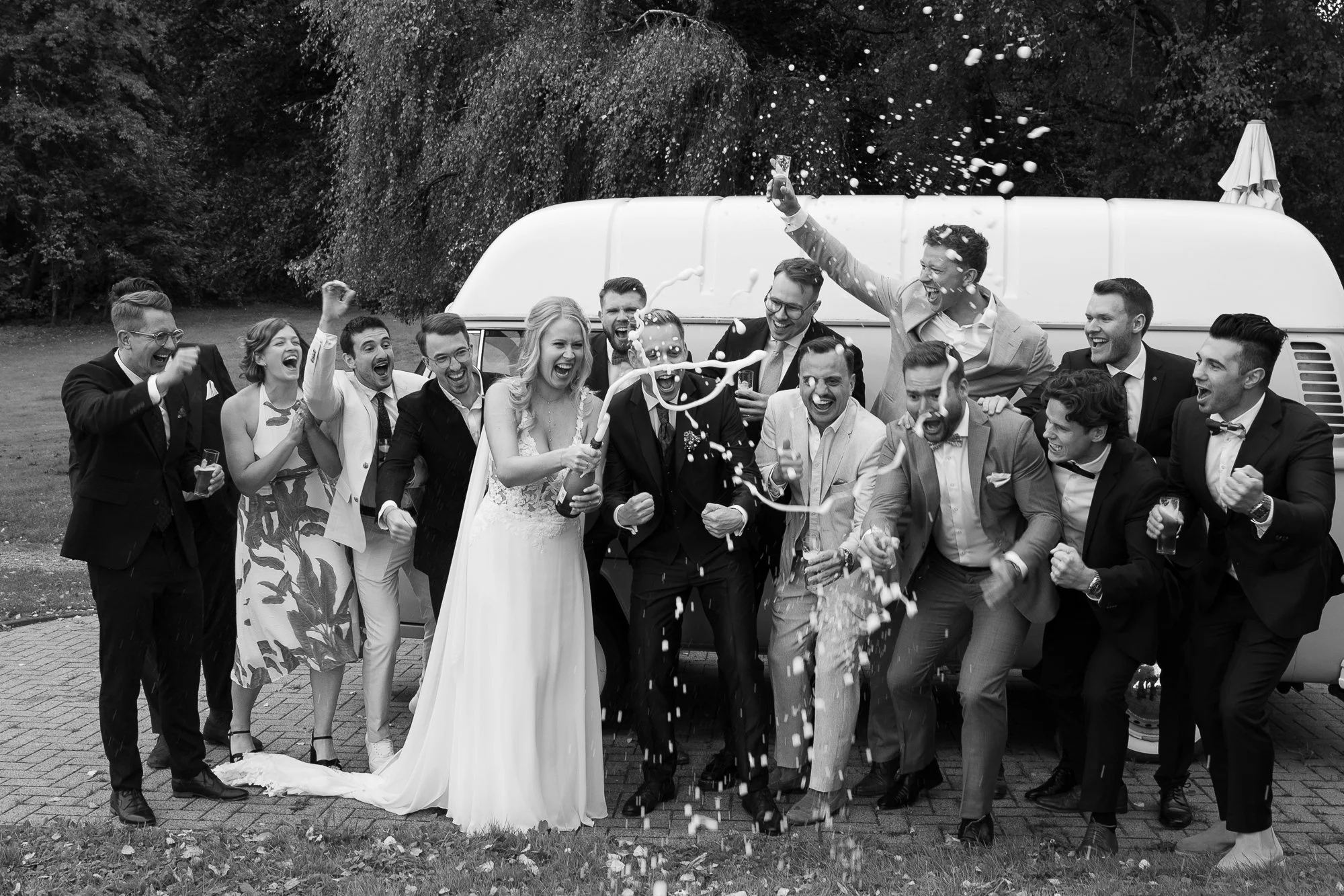 Junge Männer und Frauen in festlicher Kleidung feiern mit Sekt vor einem kleinen Bus im Freien, voller Freude, es wird Konfetti geworfen.
