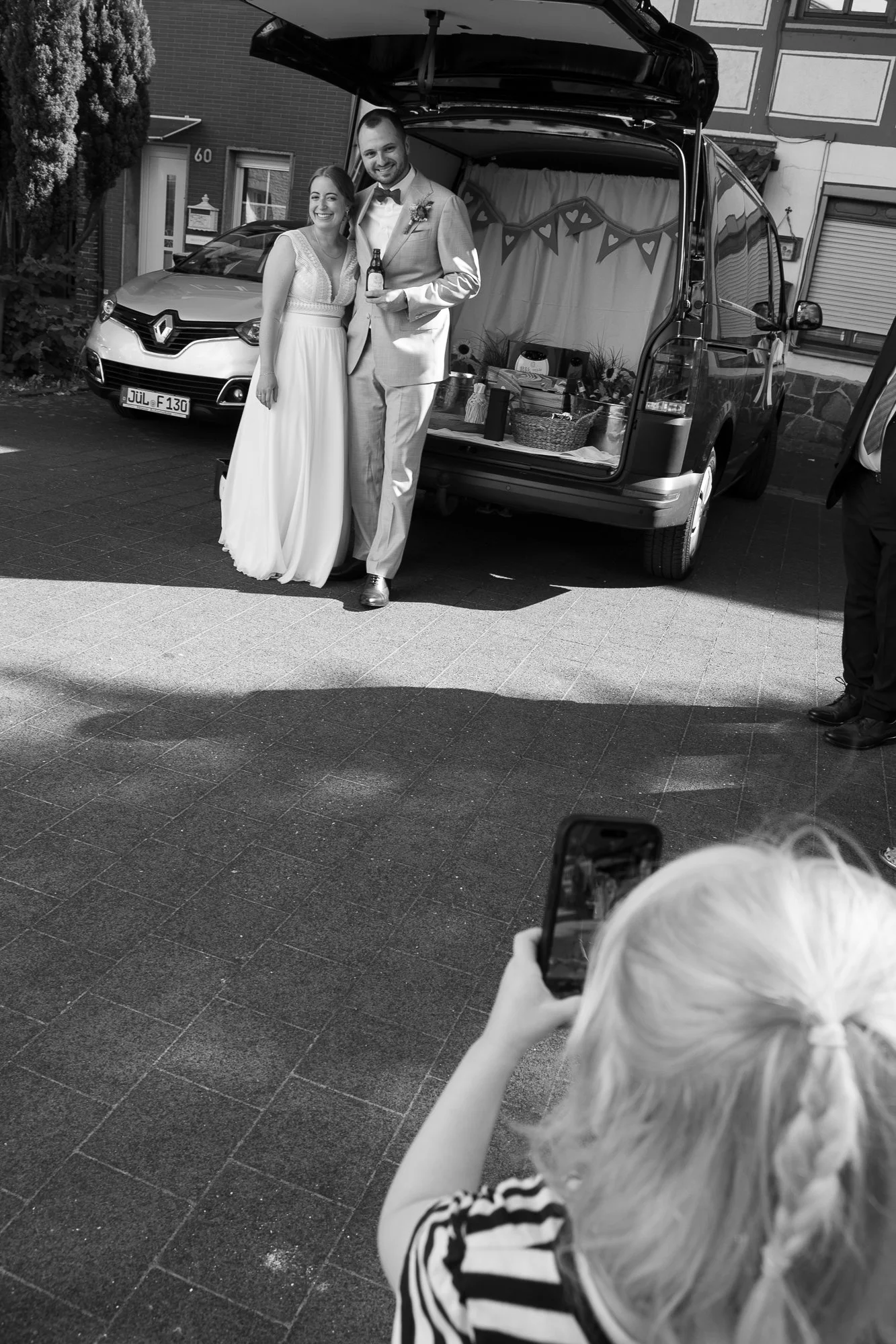 Ein glückliches Brautpaar steht vor einem umgebauten Fahrzeug bei einer Feier draußen. Die Braut trägt ein weißes Kleid, der Bräutigam einen Anzug. Eine Person im Vordergrund fotografiert das Paar mit einem Smartphone. Das Bild ist in Schwarz-Weiß.