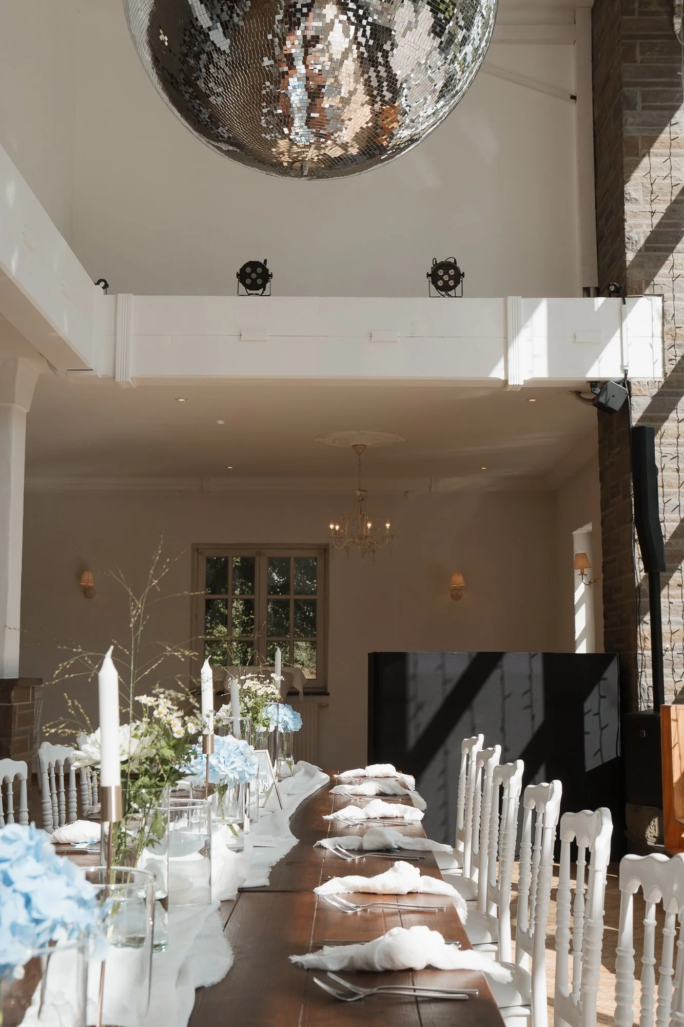 Ein festlich gedeckter langer Esstisch in einem eleganten Raum, mit weißen Stühlen, Kerzen, Blumen und Besteck, im Hintergrund ein Kronleuchter und Fenster mit Blick nach draußen.