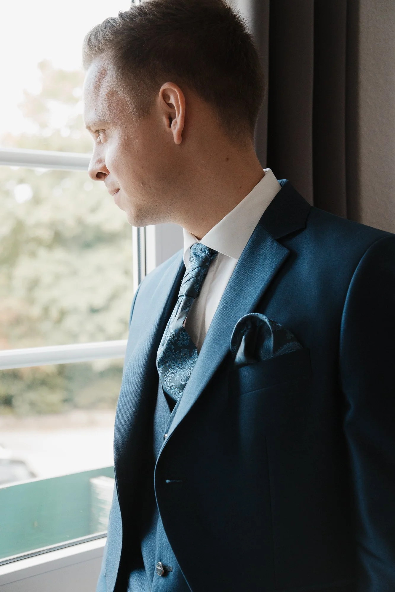 Ein junger Mann in einem dunklen Anzug und Krawatte steht neben einem Fenster und blickt nach draußen.