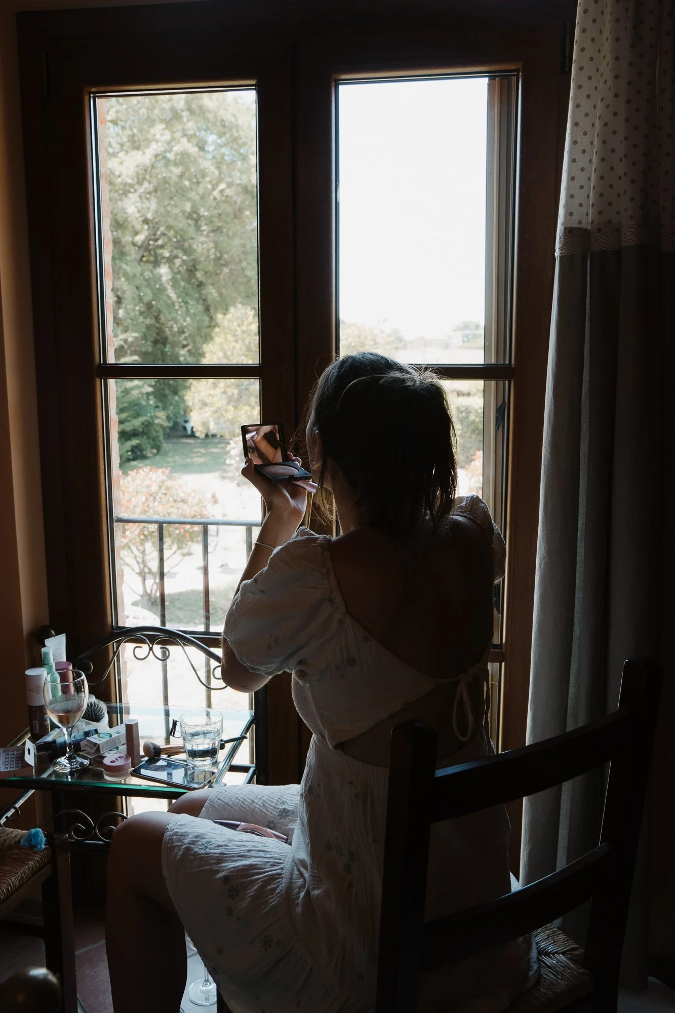 Frau sitzt vor einem Fenster, trägt ein weißes Kleid, und macht ein Selfie mit dem Handy. Am Tisch vor ihr liegen Kosmetikartikel und Gläser.