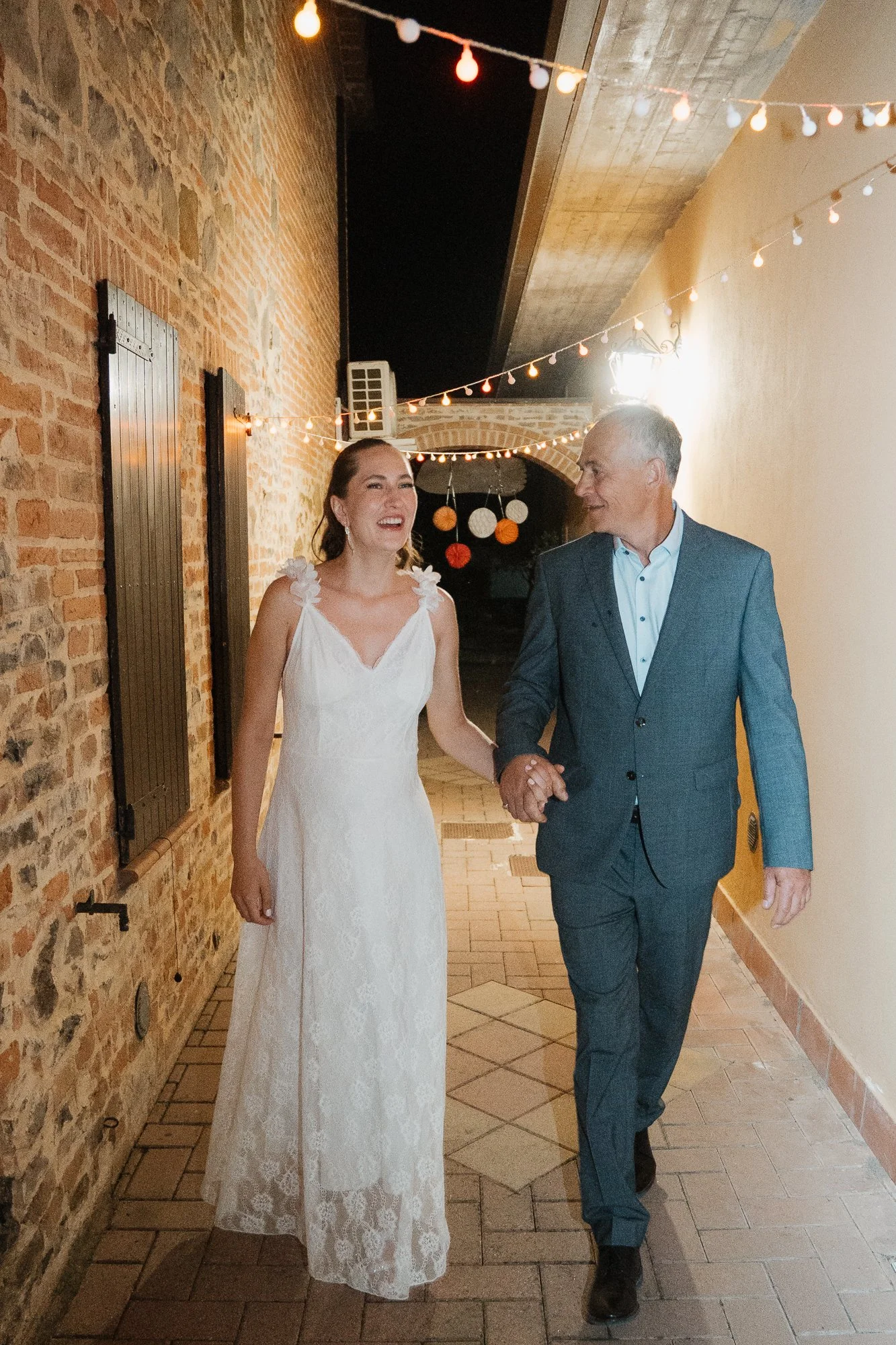 Braut in weißem Hochzeitskleid, begleitet von einem älteren Mann in Anzug, beim gemeinsamen Spaziergang. Dekorative Lichter hängen über dem Weg.