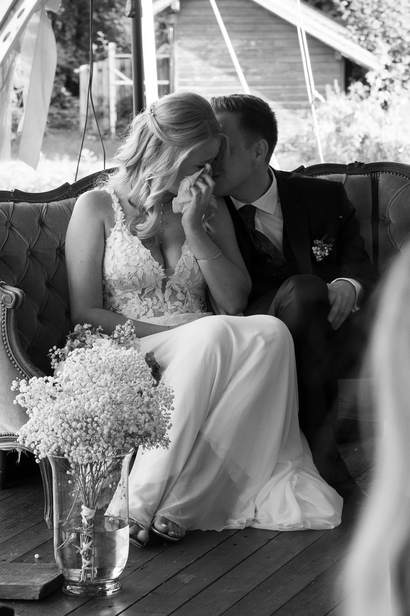 Braut und Bräutigam sitzen auf einer klassischen Couch bei einer Hochzeit, die Braut tröstet sich während der Zeremonie, neben ihnen eine Vase mit weißen Blumen.