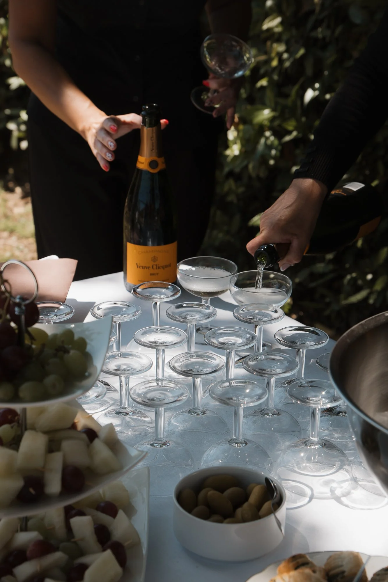 Personen servieren Sektgläser bei einer Feier im Freien. Es gibt eine Flasche Sekt, Früchte und Snacks auf einem Tisch.
